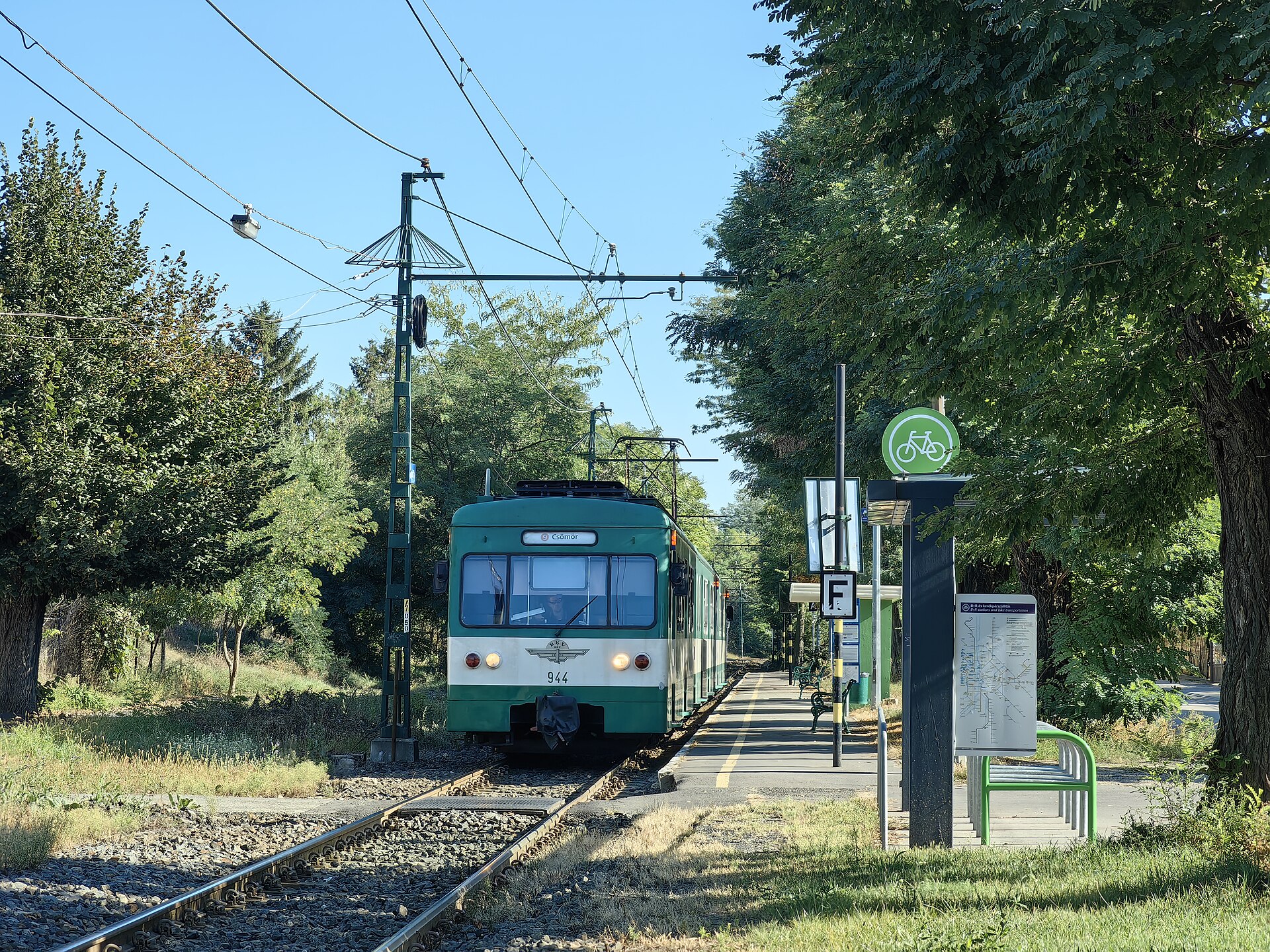 A green H9 HÉV suburban train signed for Csömör pulling into a tree-lined stop in Árpádföld, District XVI, Budapest