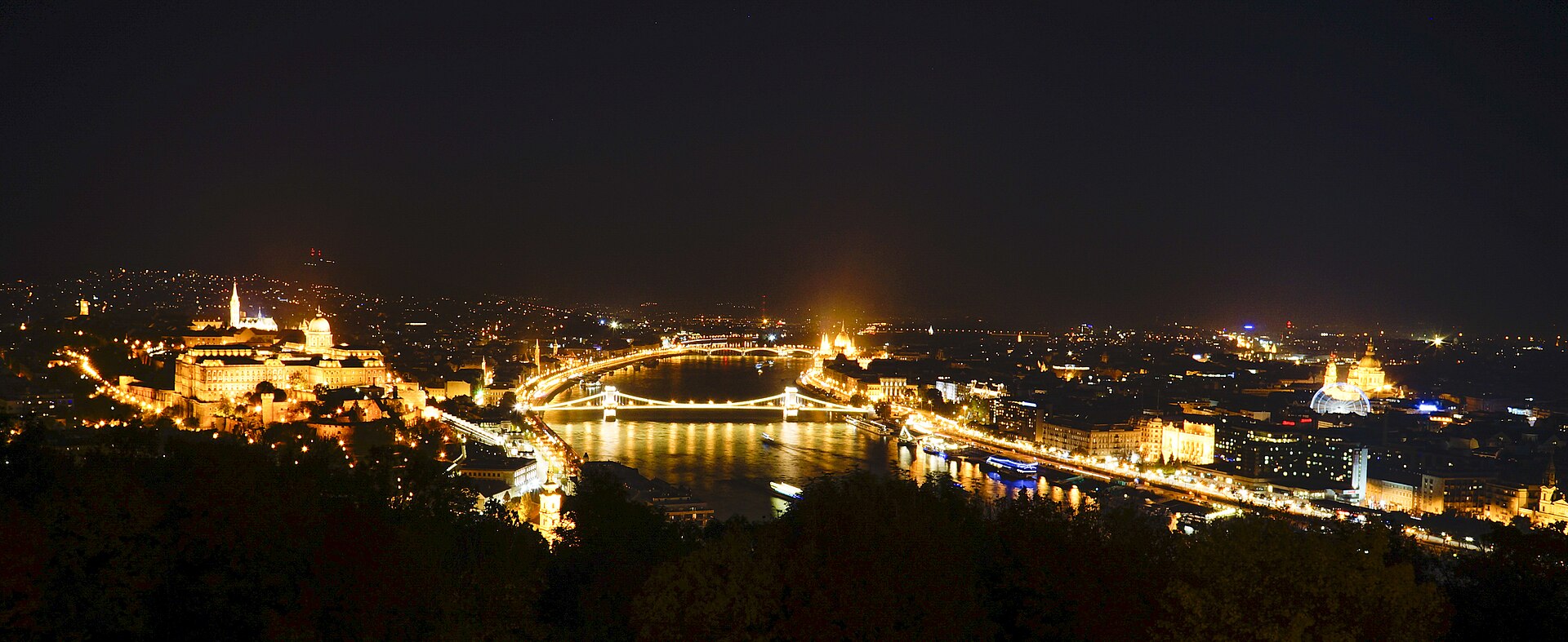 Budapest lit up at night seen from above the Danube, with Buda Castle and the Chain Bridge on the left, the Parliament dome glowing mid-frame and St Stephen’s Basilica on the right