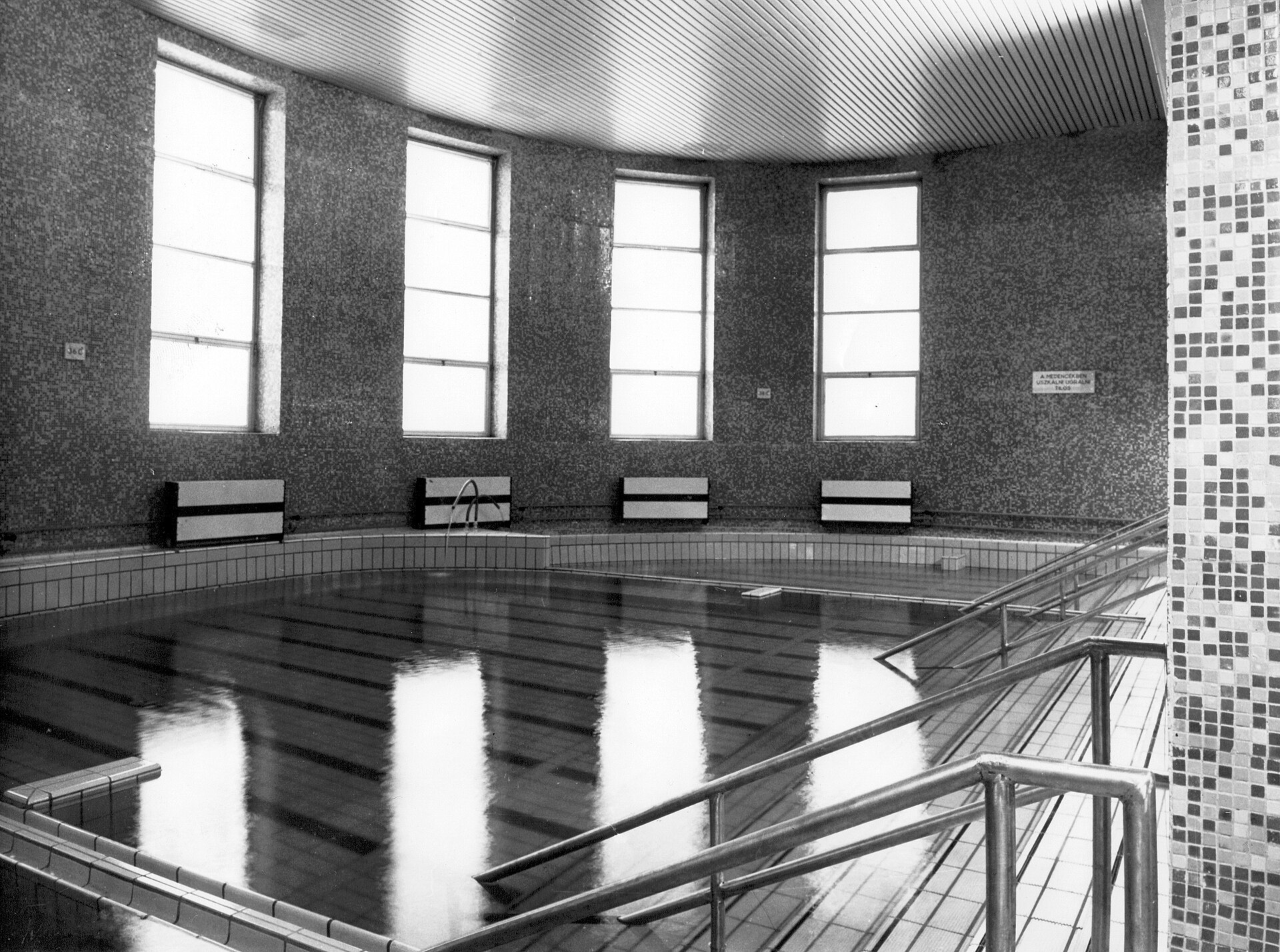 Black and white archive photo of an empty indoor thermal pool at the Dandár bath in Budapest, with tall windows, mosaic tiling and metal handrails