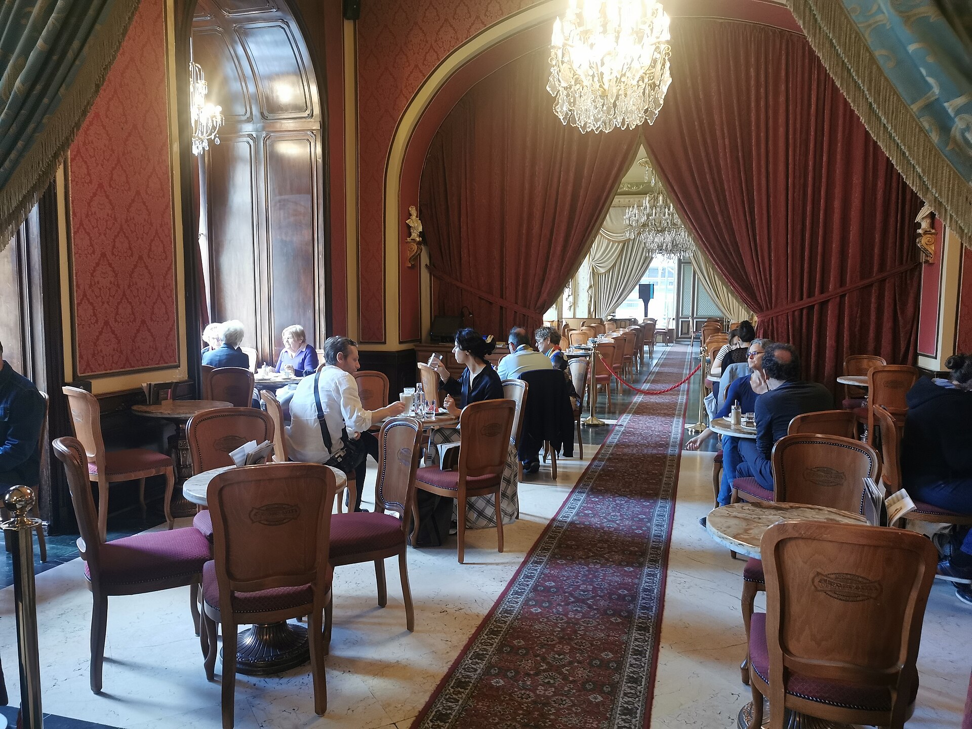 Interior of Café Gerbeaud in Budapest: guests at marble-topped tables under crystal chandeliers, with red damask walls and heavy velvet curtains