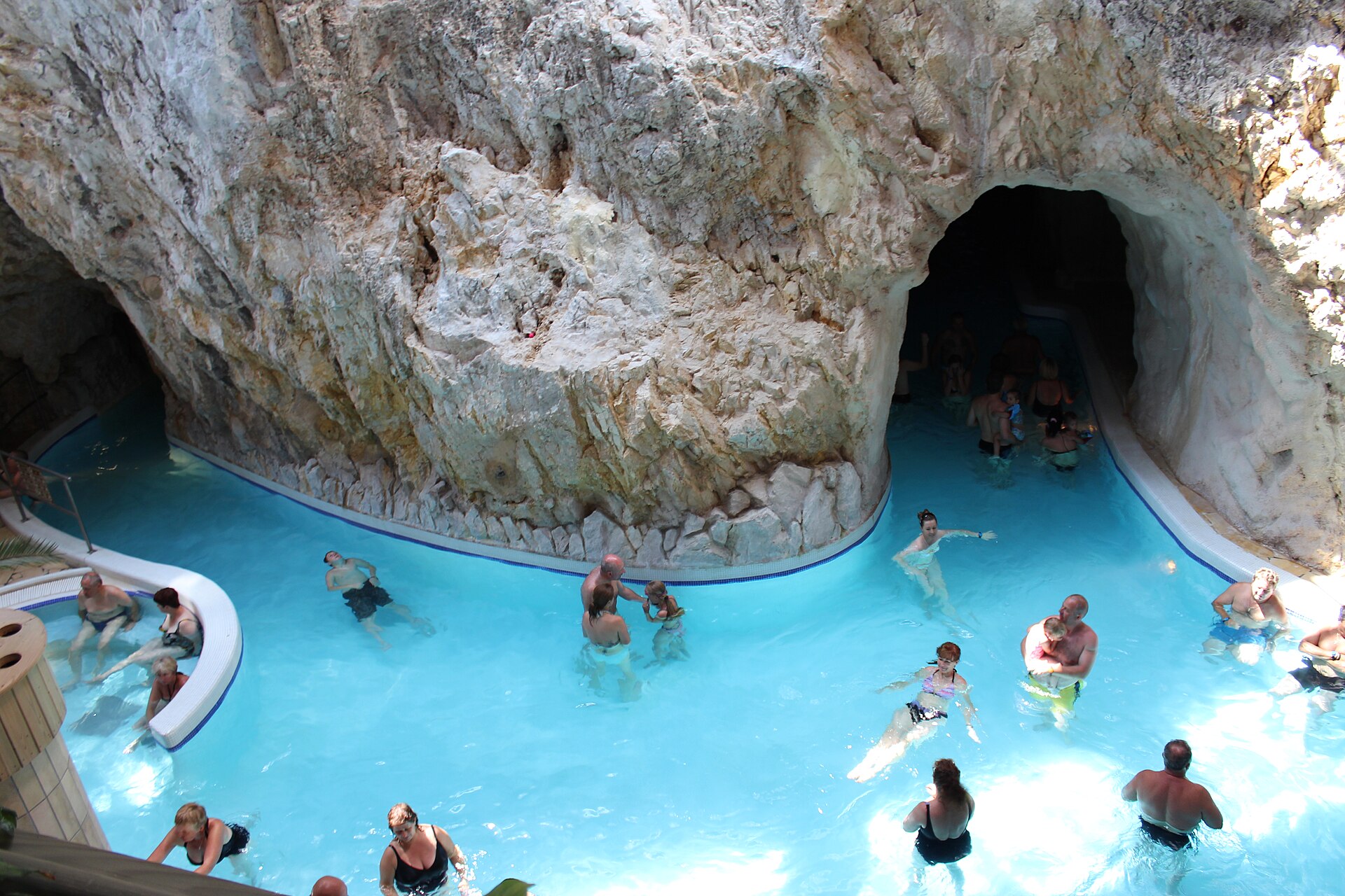 Bathers standing and swimming in bright turquoise thermal water inside the natural limestone cave pools of Miskolctapolca, with a rock archway leading to a further passage.