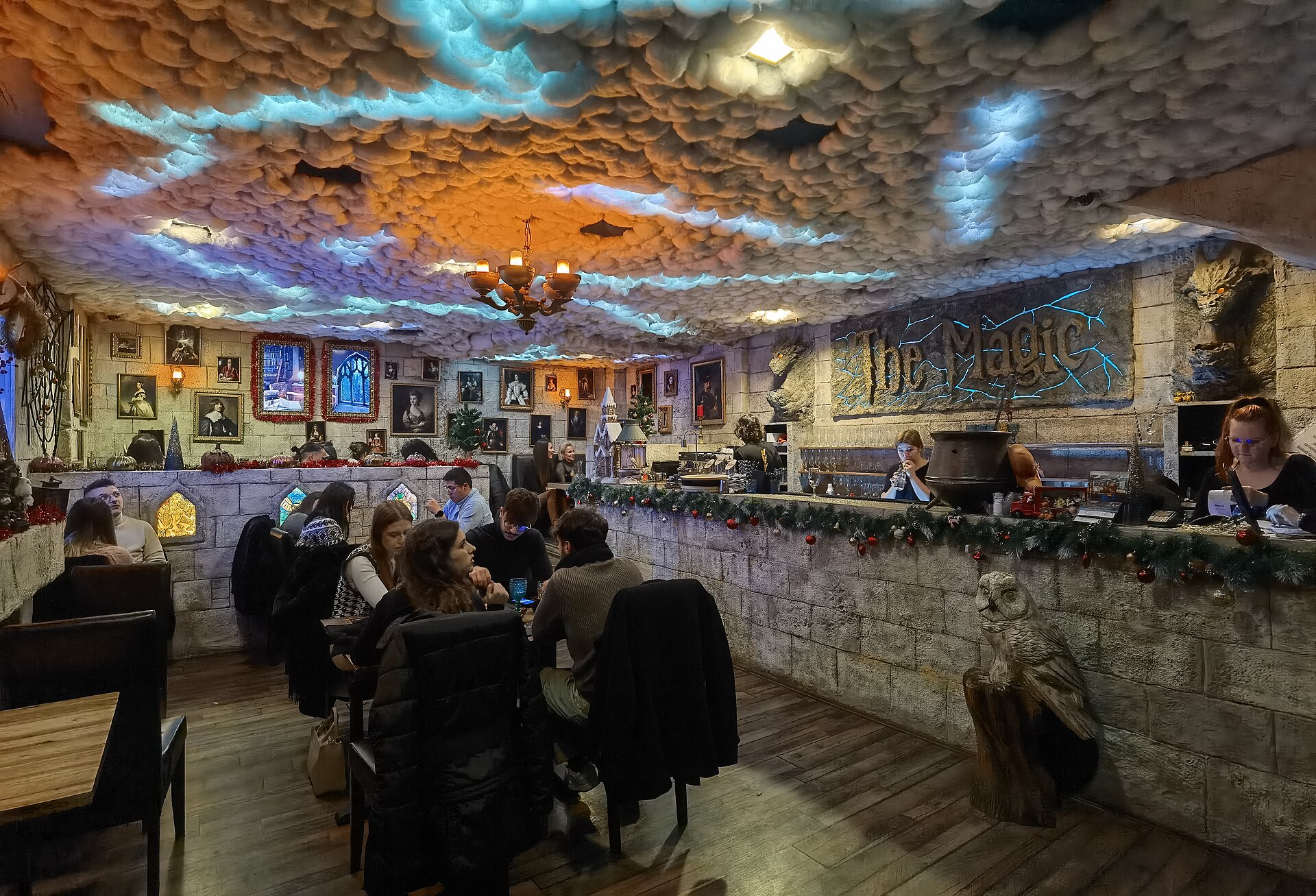 Interior of The Magic Budapest with a cloud-covered ceiling, stone walls hung with framed portraits, a carved owl and diners at wooden tables