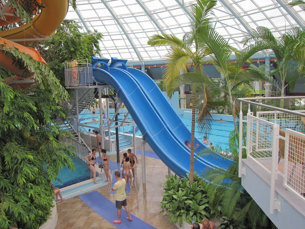 Blue twin racer slides curving down into a pool inside the glass-roofed Mediterranean Indoor Aquapark at Aquaticum Debrecen, with palm trees and swimmers around the water.