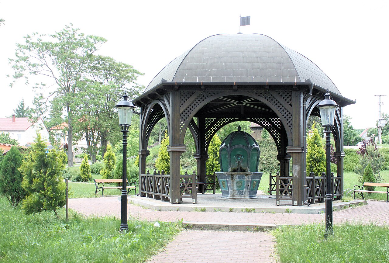 A wooden domed pavilion standing in a grassy park at Balf, sheltering a green-glazed tiled mineral spring fountain with a stone basin, framed by two cast-iron lamp posts.