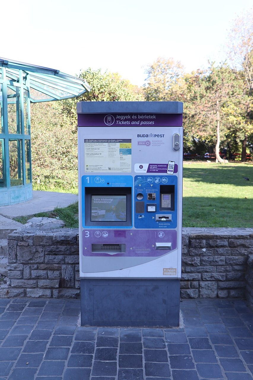 A BKK ticket machine on a paved path in a park, purple and white, headed “Jegyek és bérletek / Tickets and passes”, with a touchscreen, coin slot, card reader and banknote slot.