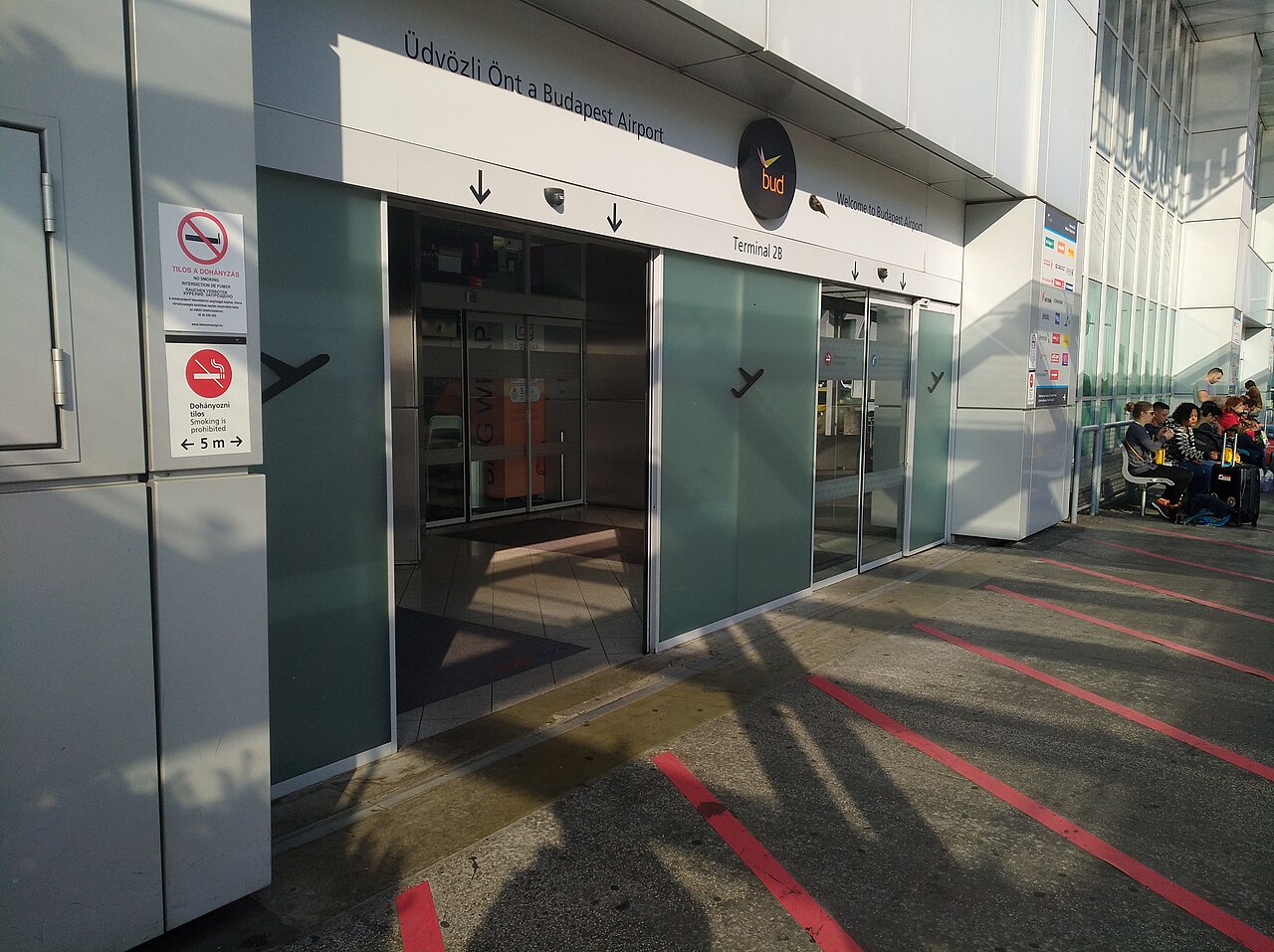 The kerbside entrance to Terminal 2B at Budapest Airport: sliding glass doors under a sign reading “Üdvözli Önt a Budapest Airport / Welcome to Budapest Airport”, with no-smoking notices on the wall and travellers waiting on benches to the right.