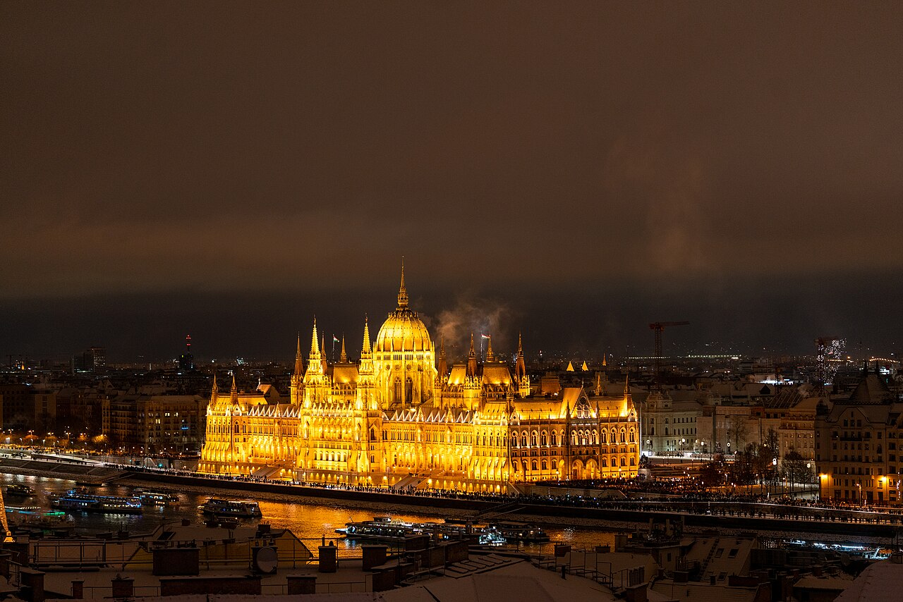 The Hungarian Parliament Building floodlit gold at night above the Danube, seen across snow-covered rooftops and chimneys, with cruise boats moored along the dark embankment below.