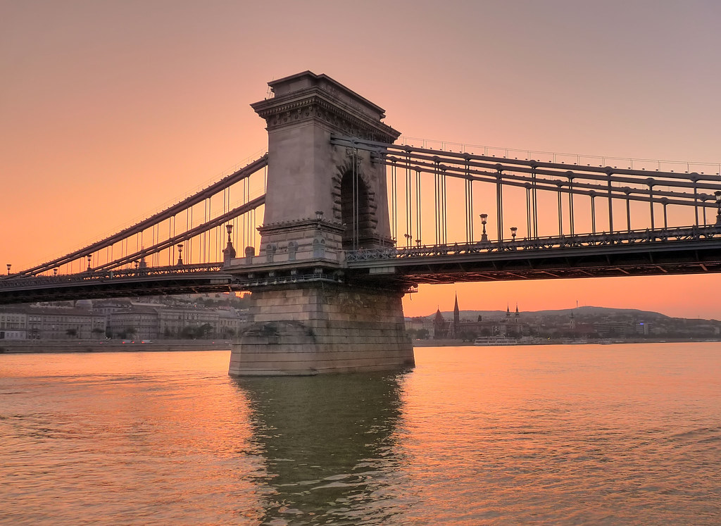 The Széchenyi Chain Bridge crossing the Danube at sunset, its stone pier rising out of still orange water, with the low Budapest skyline on the far bank.