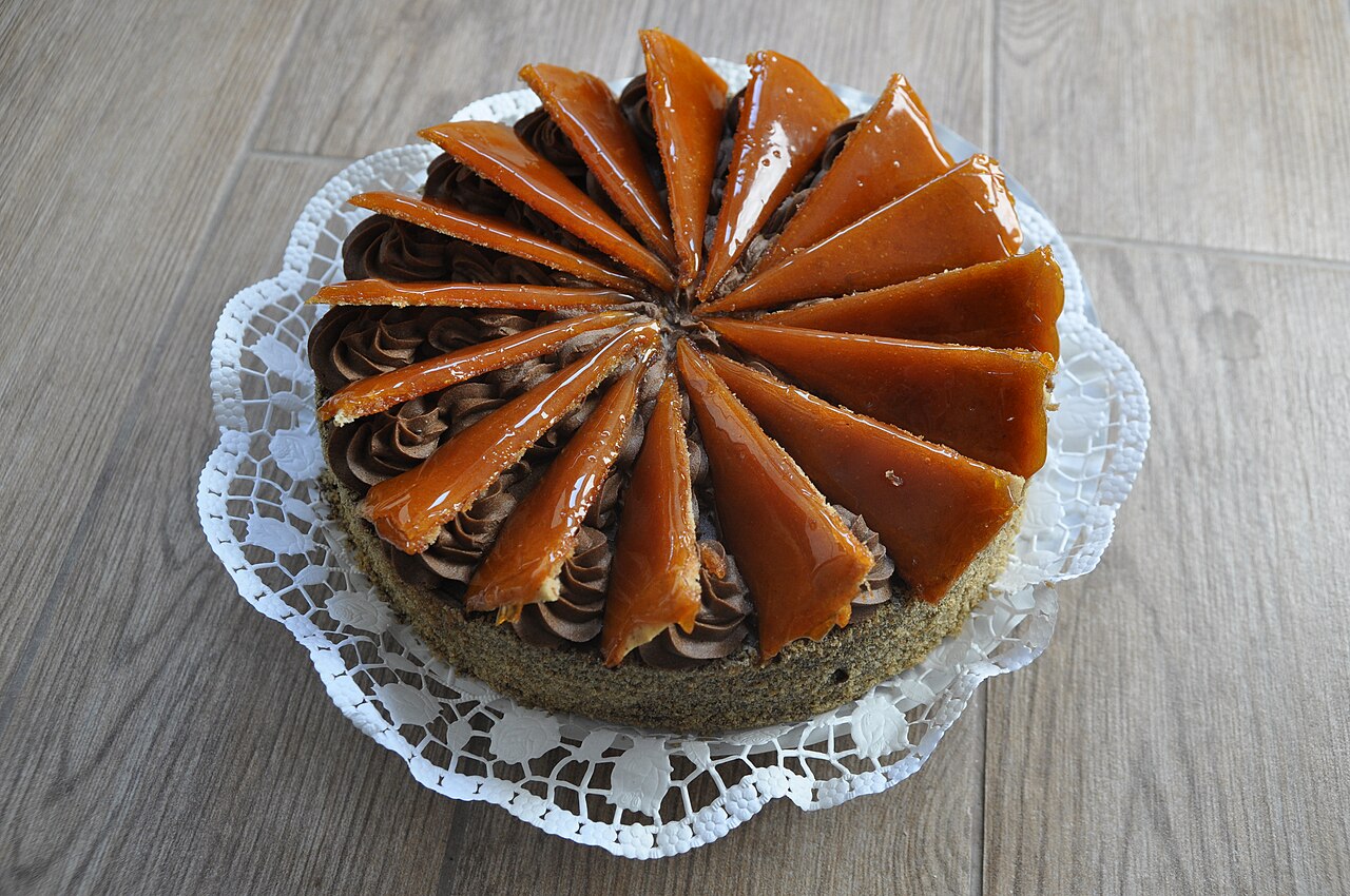 Celá Dobošova torta na bílém papírovém krajkovém tácku: piškotové vrstvy s čokoládovým máslovým krémem stříkaným po obvodu a lesklé karamelové klínky rozložené do vějíře od středu