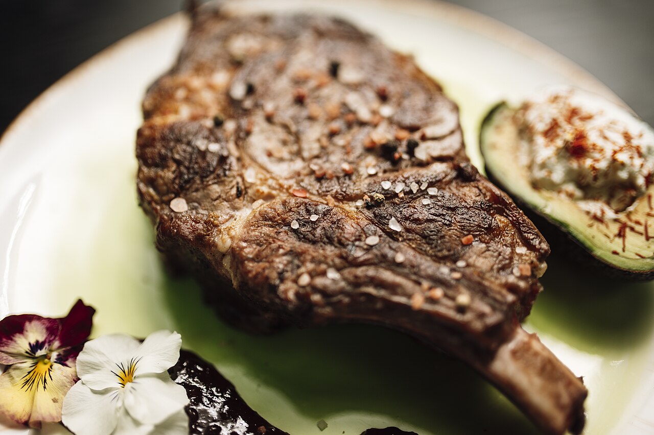 A thick bone-in steak seared dark and scattered with coarse salt and cracked pepper, plated on pale green china beside half an avocado topped with a herbed cream and fine chilli threads, with edible pansies and a dark sauce alongside.