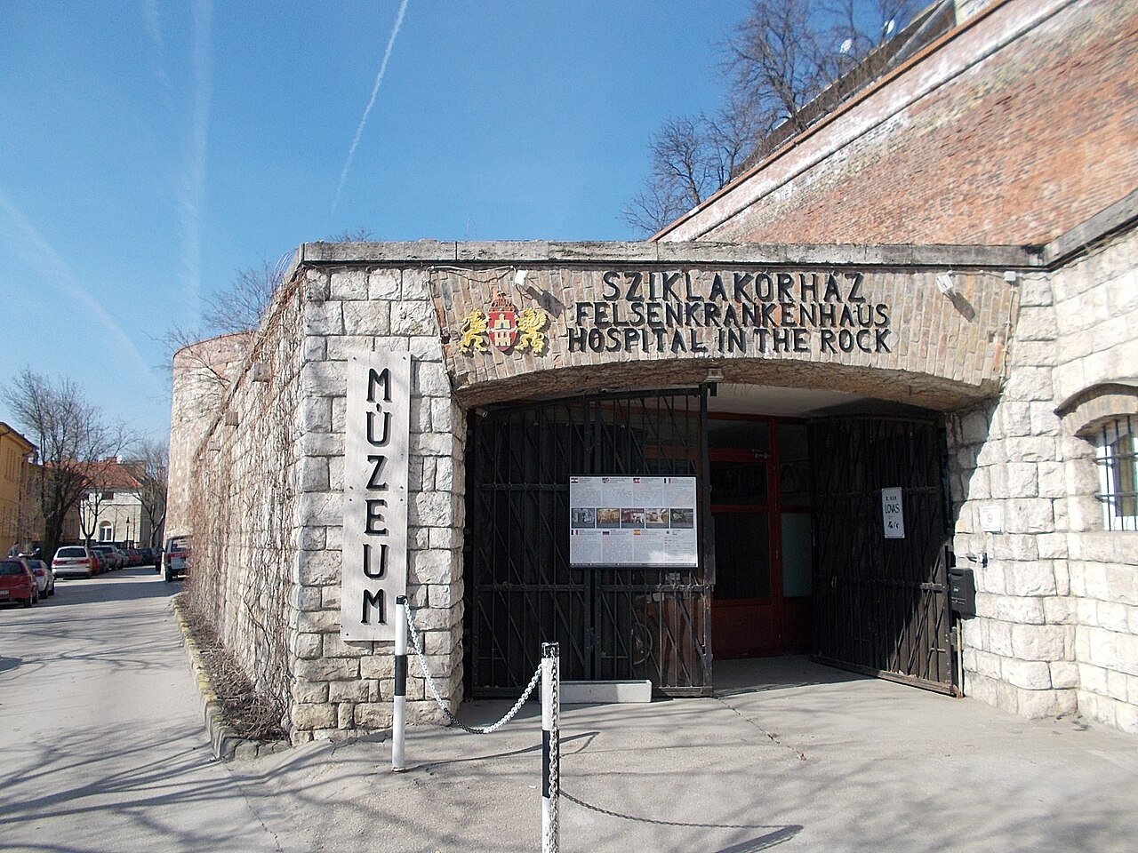 The street entrance to the Hospital in the Rock: a stone-faced portal set into a brick retaining wall, with iron gates, a vertical MÚZEUM sign and lettering reading SZIKLAKÓRHÁZ, FELSENKRANKENHAUS and HOSPITAL IN THE ROCK above the arch.