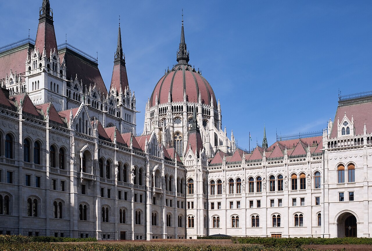 The Hungarian Parliament Building from ground level: a white neo-Gothic facade of pointed arches, pinnacles and spires under a red-tiled central dome, with clipped hedges along the base and a Hungarian flag on the roofline.