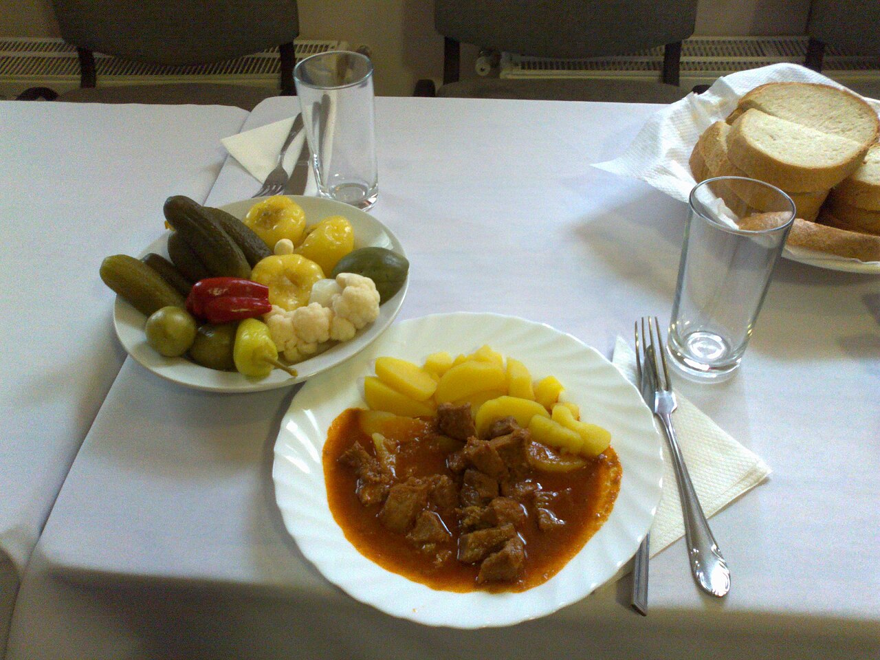 A white plate of Hungarian pörkölt, cubes of meat in thick red paprika gravy, served with boiled potatoes, beside a dish of preserved gherkins, wax peppers and cauliflower and a basket of sliced white bread.
