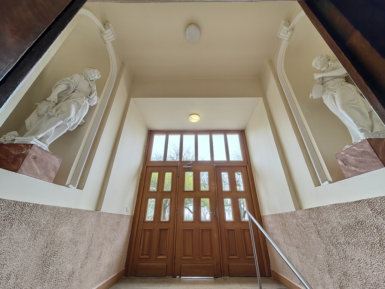 The covered entrance porch of Lukács Bath seen from the foot of the steps: two white robed statues standing in tall wall niches either side of wooden double doors with glazed panels.