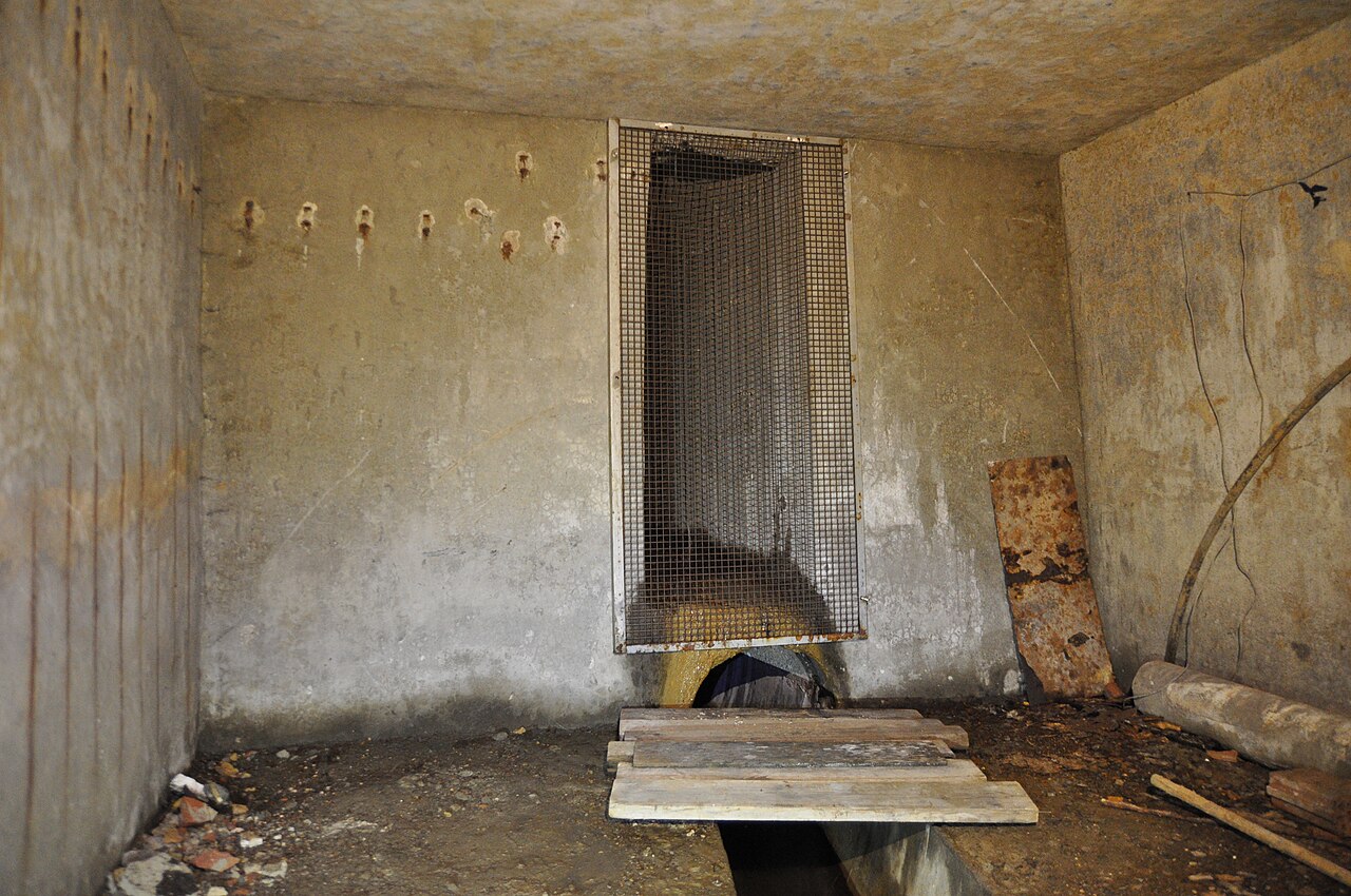 A bare concrete chamber housing a thermal spring outlet: water emerging from a grated opening in the wall into a channel cut across the floor, with rough planks laid over it.