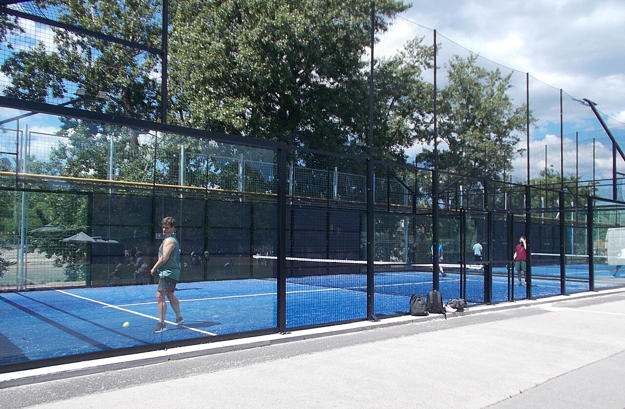 A man in a green vest mid-rally on a blue padel court enclosed by glass panels and black mesh, with a second court and more players behind him, leafy trees overhead and backpacks left on the kerb outside.