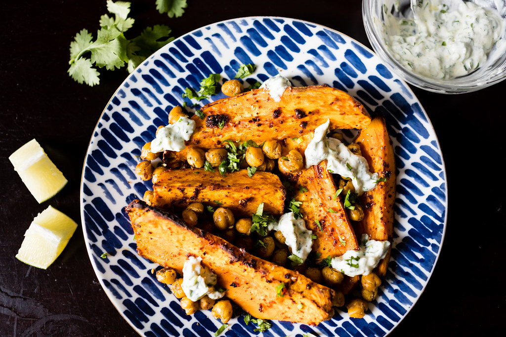 Roasted sweet potato wedges with spiced chickpeas, chopped coriander and a herbed white dressing on a blue-patterned plate, with lemon wedges alongside.