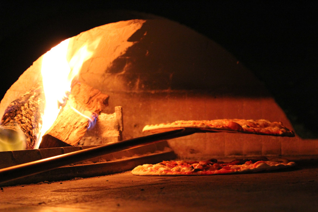 Două pizza subțiri care se coc pe vatra de piatră a unui cuptor cu lemne boltit, cu lopata lungă de metal întinsă înăuntru și buștenii arzând în spate