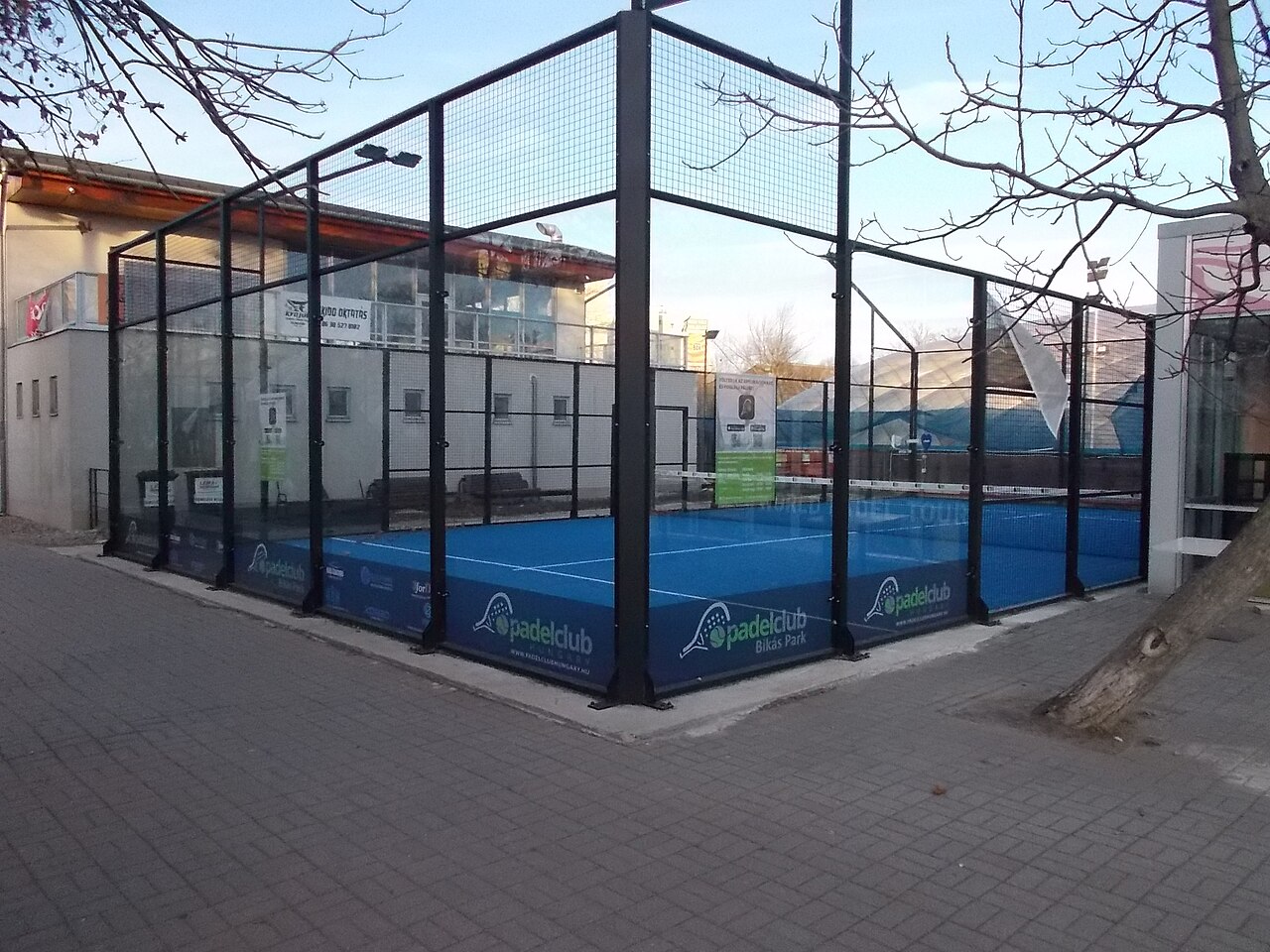 Ein leerer Padelplatz mit blauem Kunstrasen, Glasseitenwänden und schwarzem Stahlgitterrahmen, aufgestellt auf einem gepflasterten Platz zwischen kahlen Winterbäumen, mit dem Schriftzug padelclub Bikás Park auf den Bodenbanden