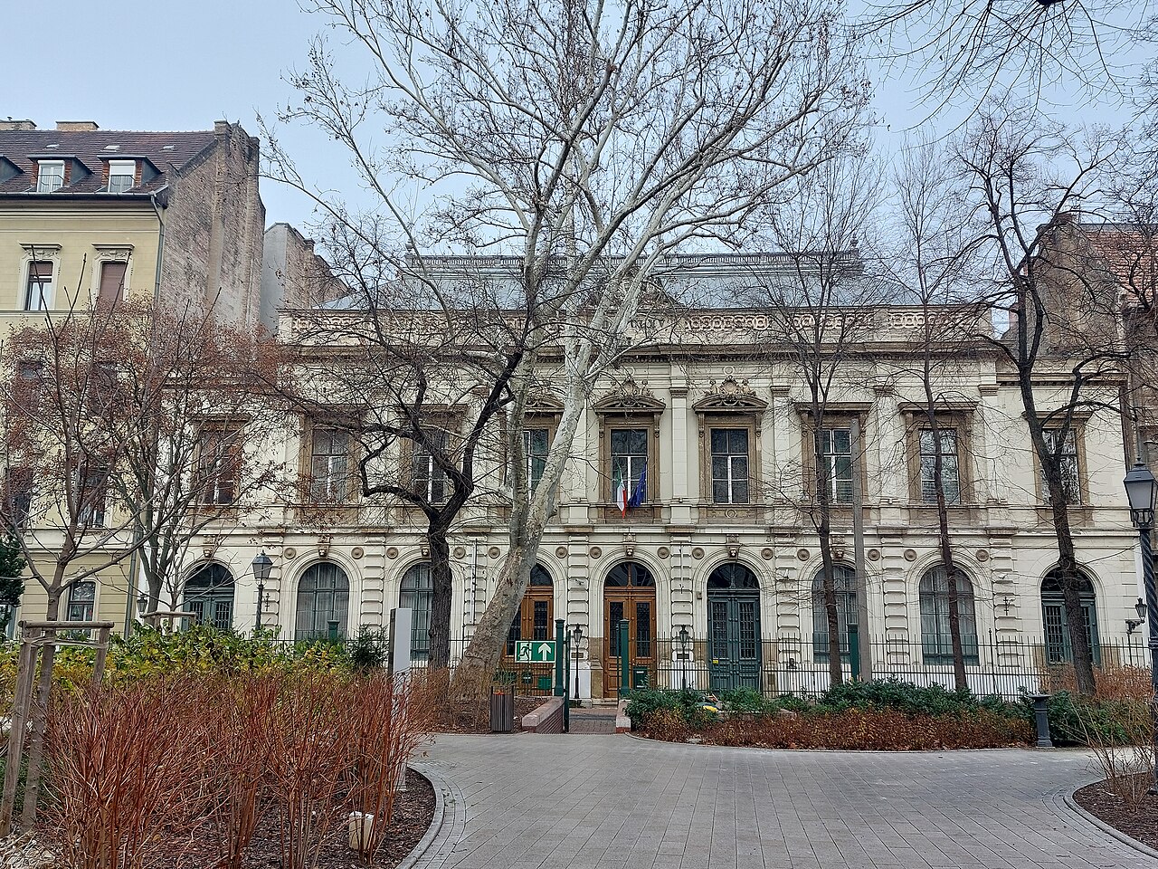 L’ancienne Chambre des représentants sur Bródy Sándor utca : un long bâtiment néo-Renaissance de deux étages en pierre claire, des fenêtres cintrées au rez-de-chaussée derrière des grilles de fer, les drapeaux italien et hongrois au-dessus de la porte centrale, et des platanes d’hiver dénudés sur la place pavée devant.