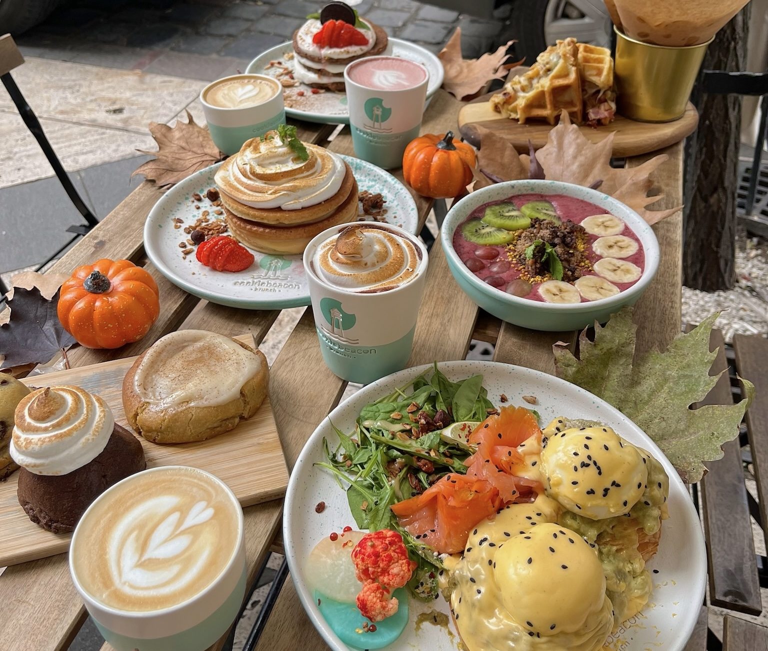 An autumn brunch table on a Cookie Beacon pavement terrace: eggs Benedict with smoked salmon, meringue-topped pancakes, a berry smoothie bowl, cookies and coffees