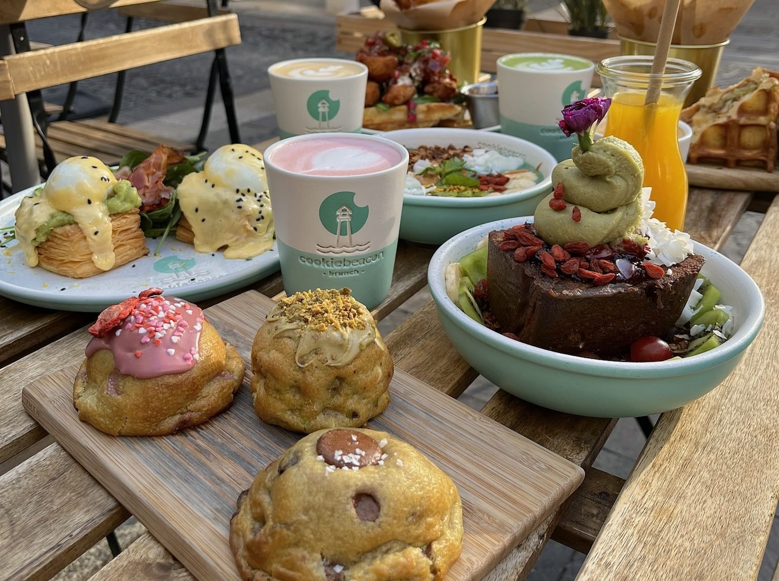 Tres cookies gruesas sobre una tabla en el local de la Basílica de Cookie Beacon, con huevos Benedict, un latte rosa y un bol de bizcocho de chocolate detrás