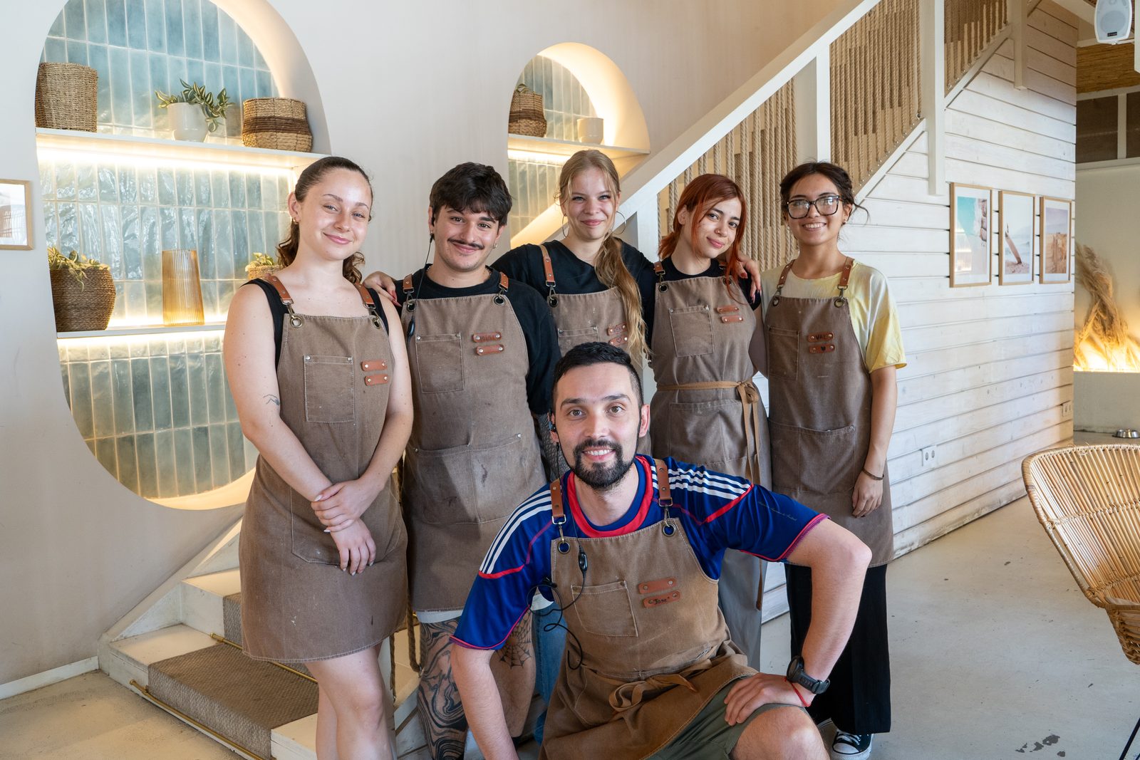 Six Cookie Beacon staff in brown canvas aprons in front of lit tiled alcoves and a white staircase