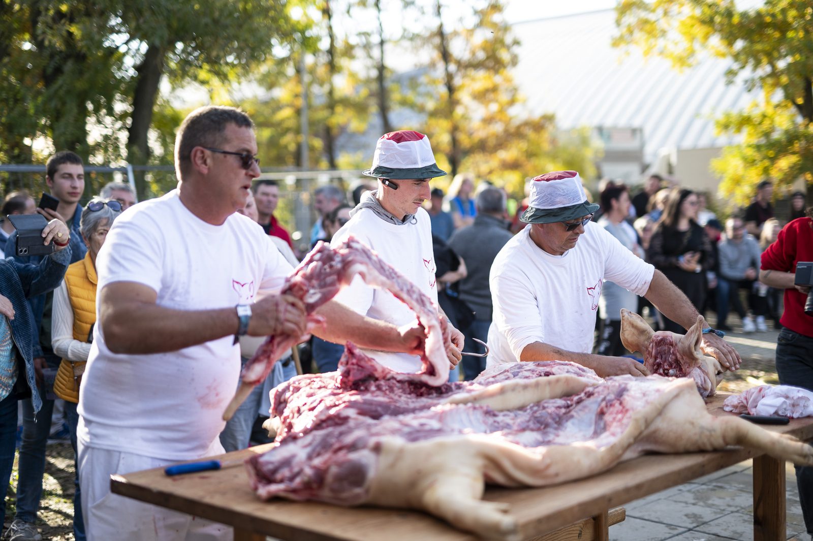 Three men in white T-shirts cutting up a pig carcass on a wooden table outdoors, onlookers filming behind them
