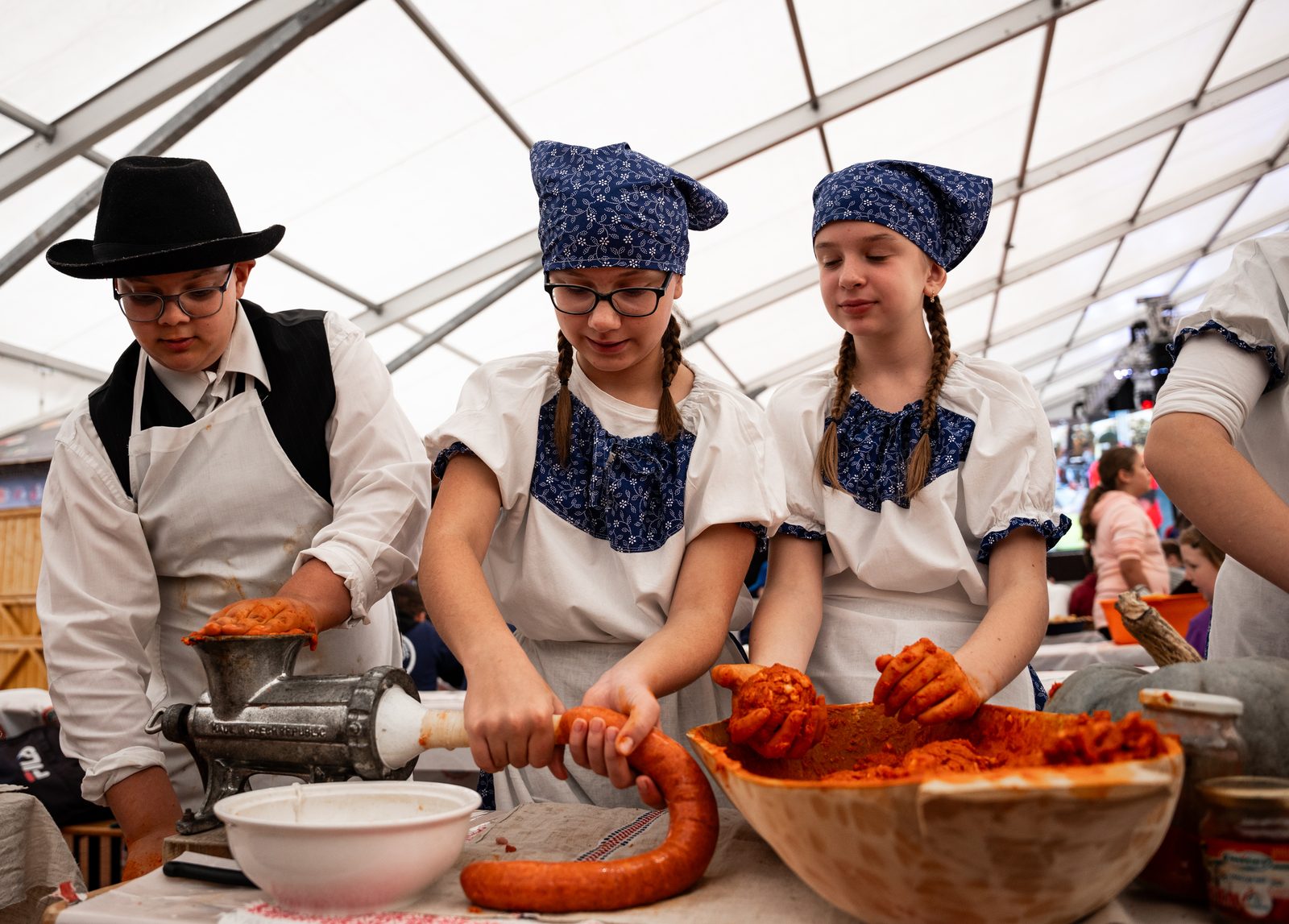 Children at a contest table, a boy in a black hat, black waistcoat, white shirt and apron feeding a hand grinder, two girls in blue-and-white folk costume beside him, one easing a long red sausage off the stuffer, the other mixing spiced meat in a wooden bowl