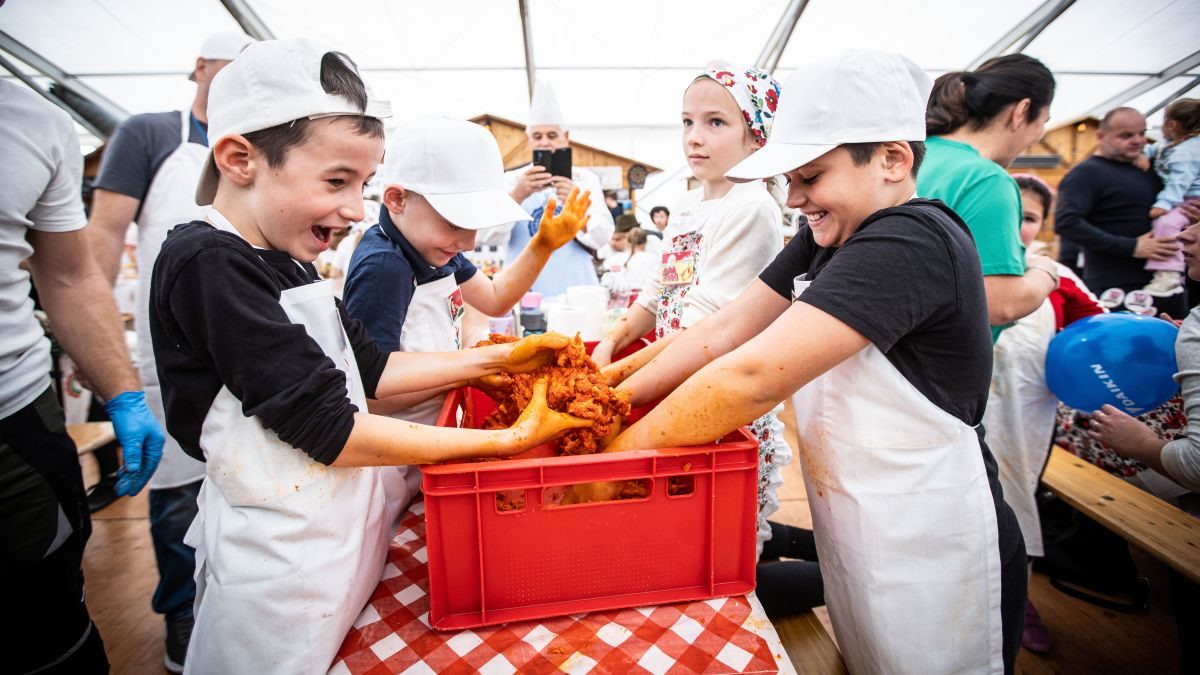 Children in white aprons and caps kneading paprika-red sausage meat in a red crate on a checked tablecloth