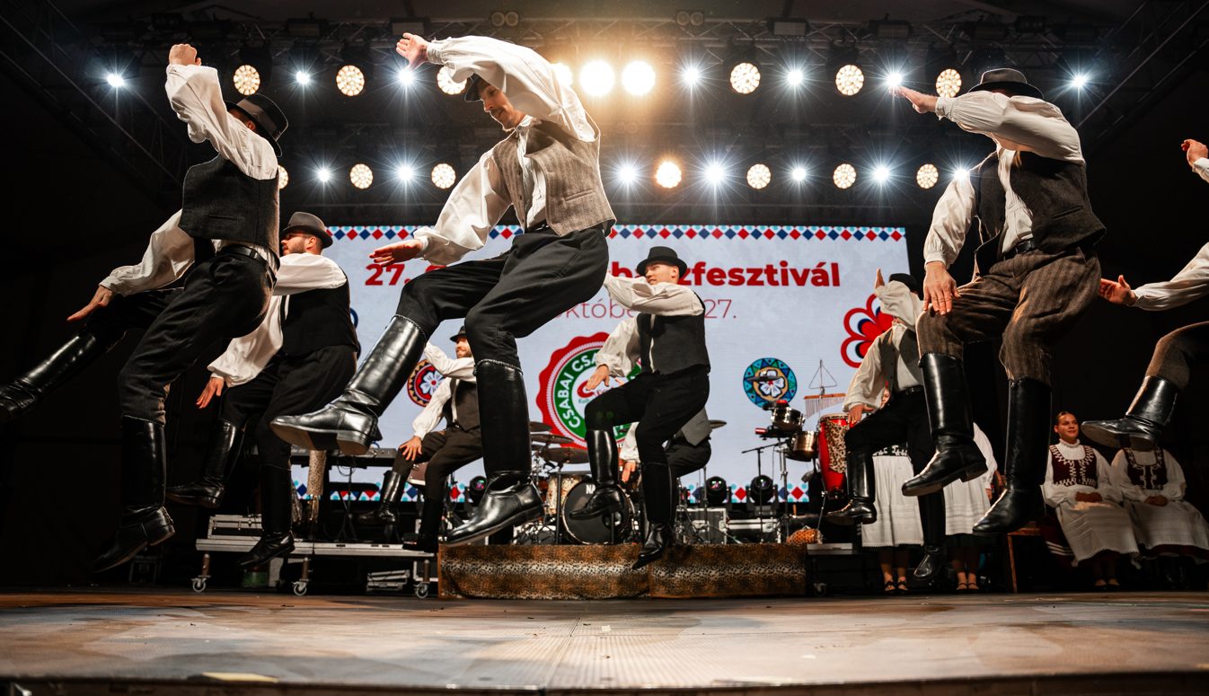Male folk dancers in black boots, waistcoats and hats leaping mid-air on a lit stage, the festival banner behind them