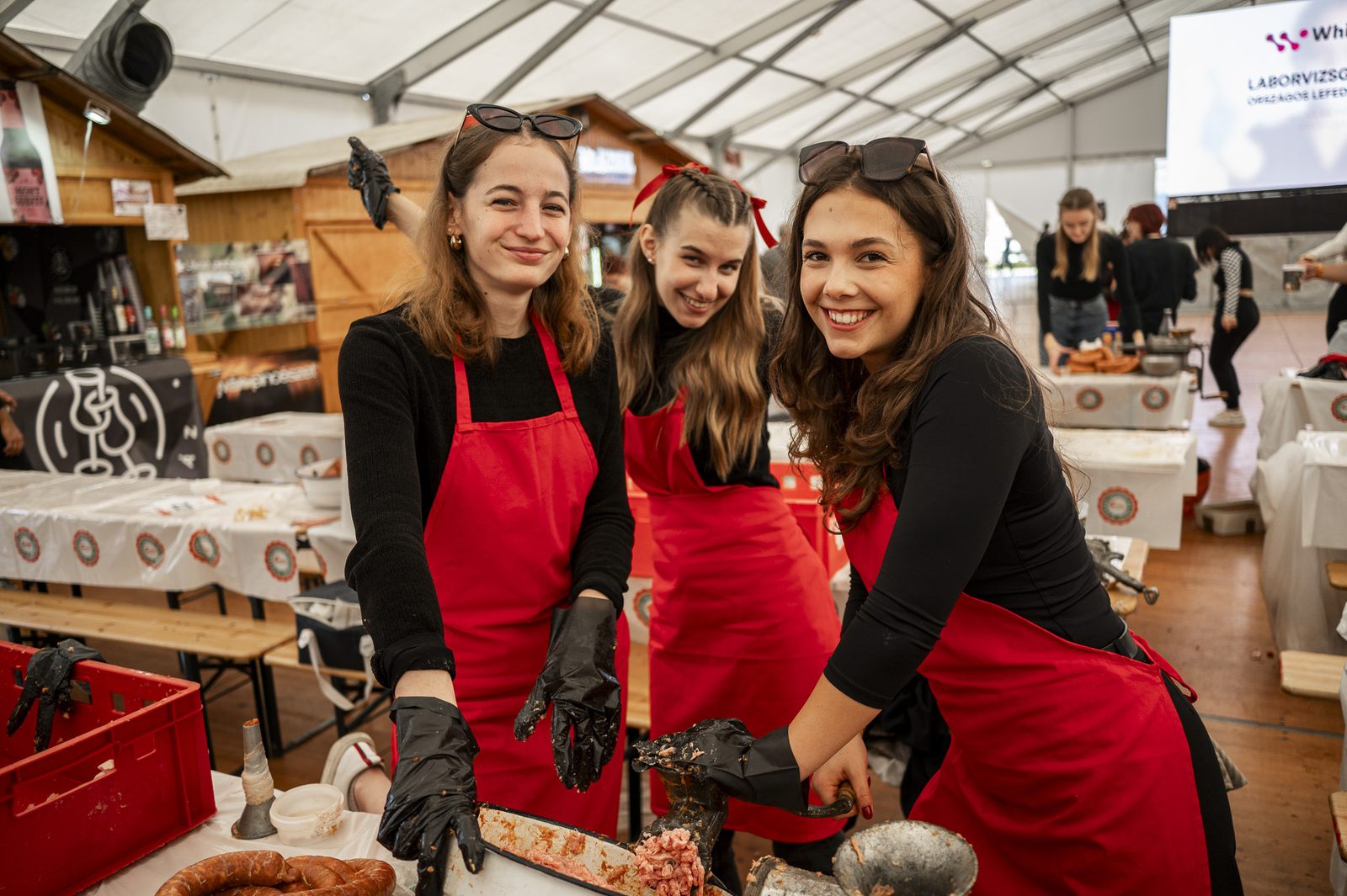 Three young women in red aprons and black gloves at a hand grinder, a bowl of minced meat and a coil of sausage in front of them