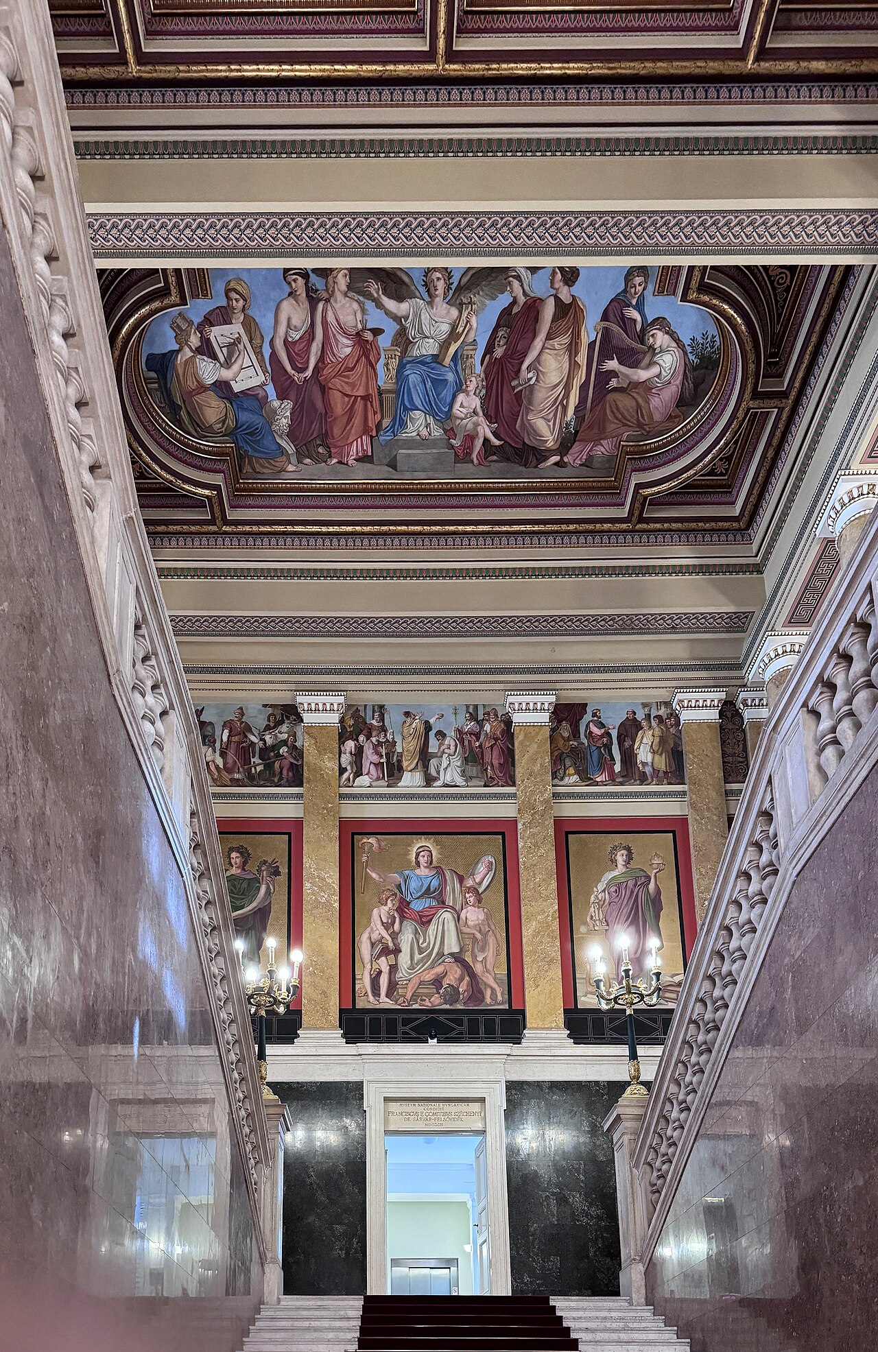 La escalinata principal del Museo Nacional Húngaro vista desde abajo: peldaños de mármol entre dos balaustradas, paneles alegóricos pintados sobre pared roja, techo artesonado dorado y una inscripción fundacional en latín sobre la puerta del fondo.