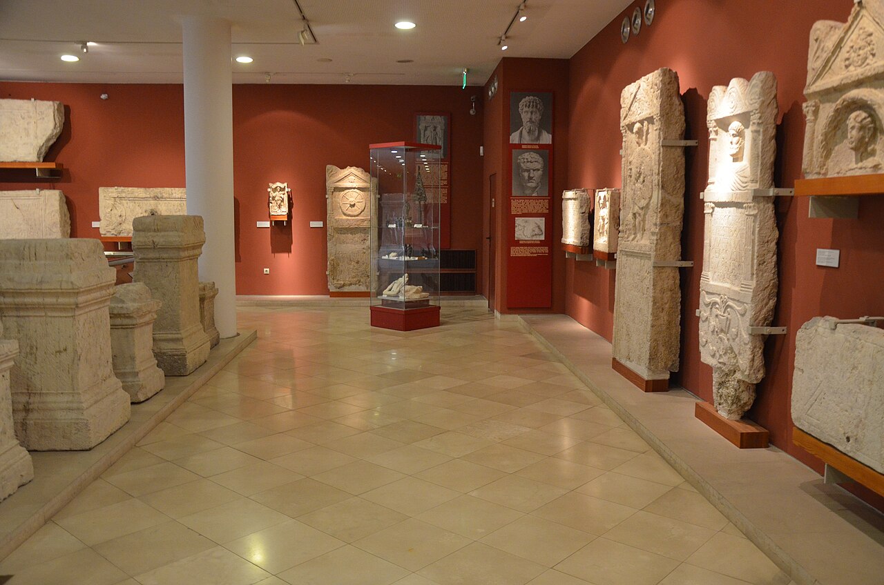 El lapidario romano del Museo Nacional Húngaro: estelas funerarias y altares de piedra caliza montados a lo largo de paredes rojo oscuro, con una vitrina en el centro de una galería de suelo embaldosado.
