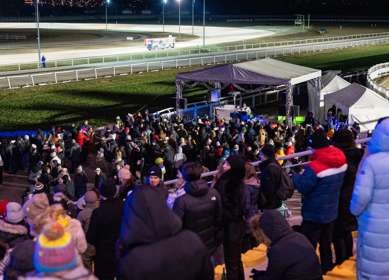 Público con abrigos y gorros de invierno en los escalones de la tribuna alrededor de una carpa de DJ en Kincsem Park de noche, con la pista iluminada al fondo