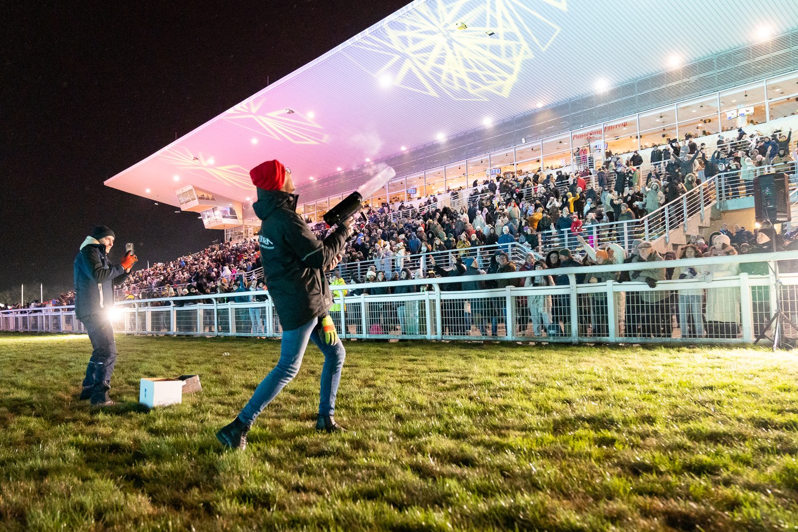 Un hombre con gorro rojo apunta un cañón de confeti hacia la tribuna llena de Kincsem Park de noche, y sobre las gradas el piso acristalado con el letrero Panoráma Étterem