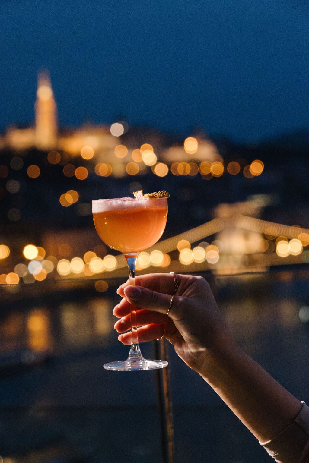 A hand raising a pink cocktail against the lit Chain Bridge and Buda Castle at night from the Liz & Chain terrace