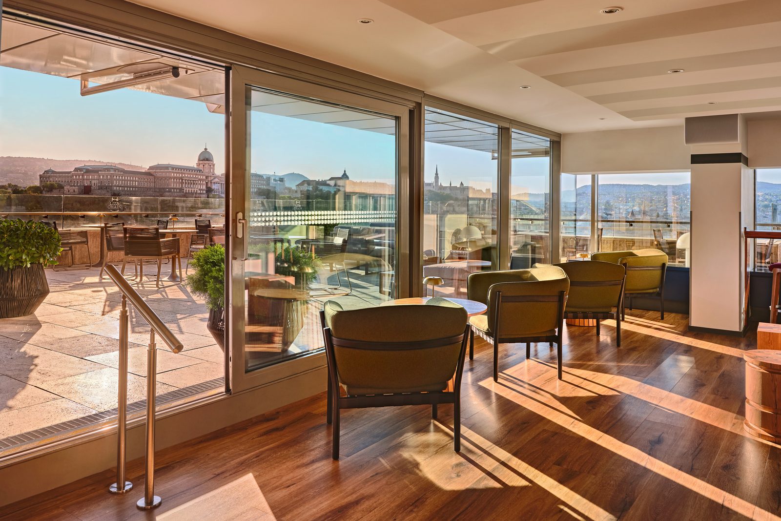 The indoor lounge of Liz & Chain in daylight, floor-to-ceiling glass, armchairs and low tables, with Buda Castle in the window