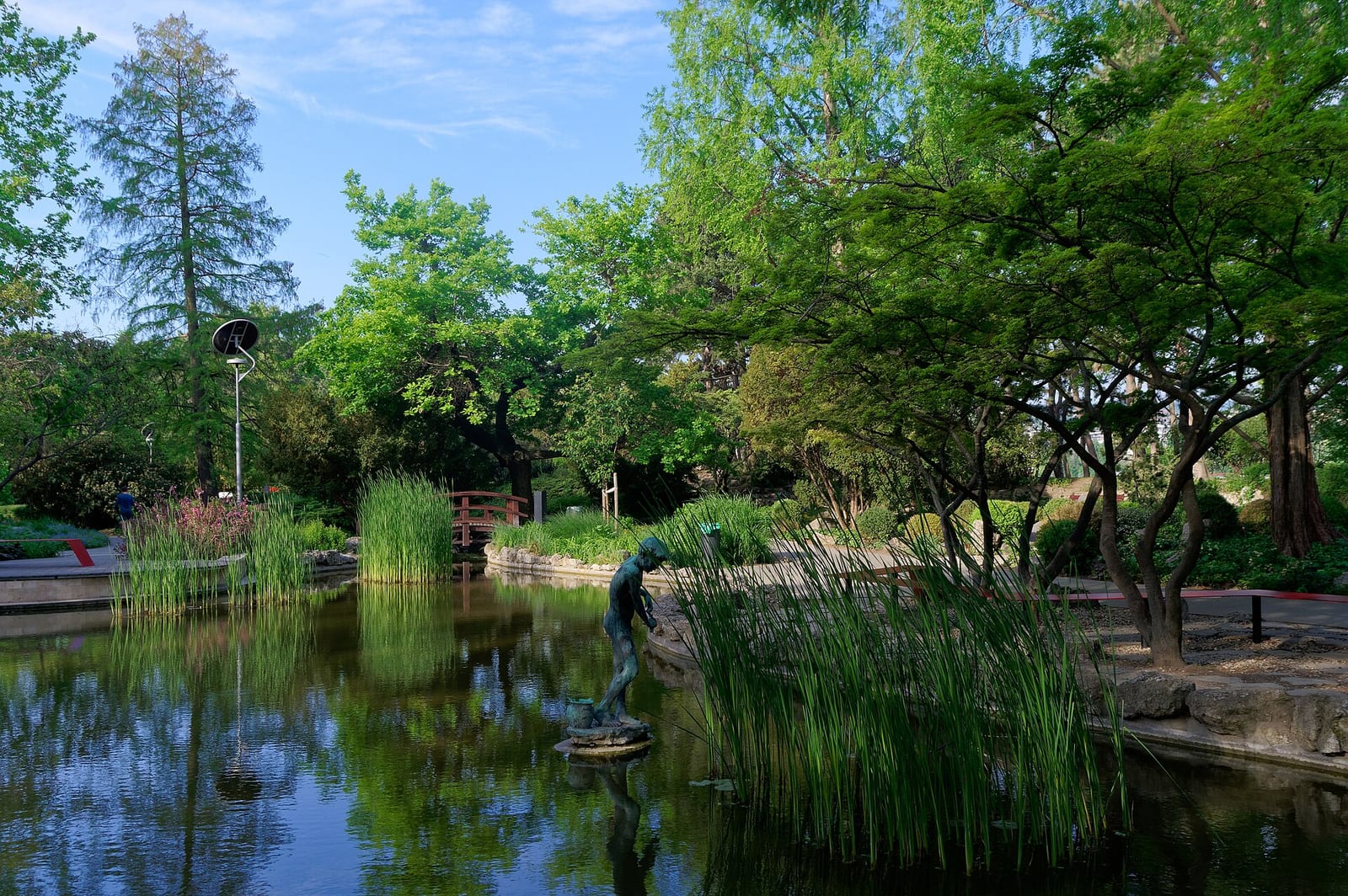 L’étang du jardin japonais de l’île Marguerite, avec des roseaux, une petite statue et un pont de bois rouge à l’arrière-plan