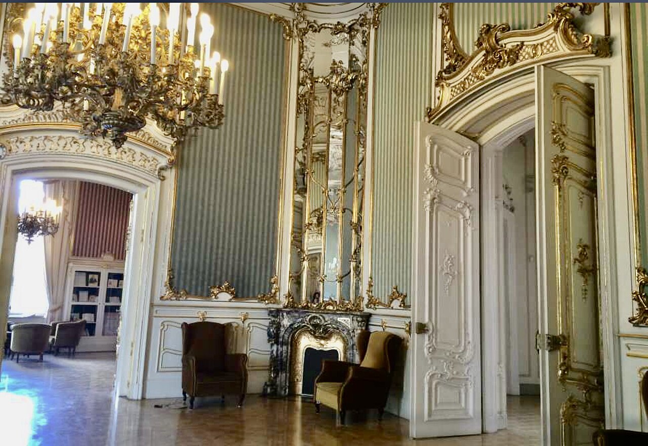 Un salon du palais Wenckheim : des panneaux muraux rayés bleu-vert pâle bordés de rinceaux dorés, un haut lustre à bougies doré, une cheminée de marbre sous un trumeau à miroir, deux fauteuils à côté, une double porte blanche à ornements dorés restée ouverte, et une autre pièce garnie de rayonnages visible par l’arcade.