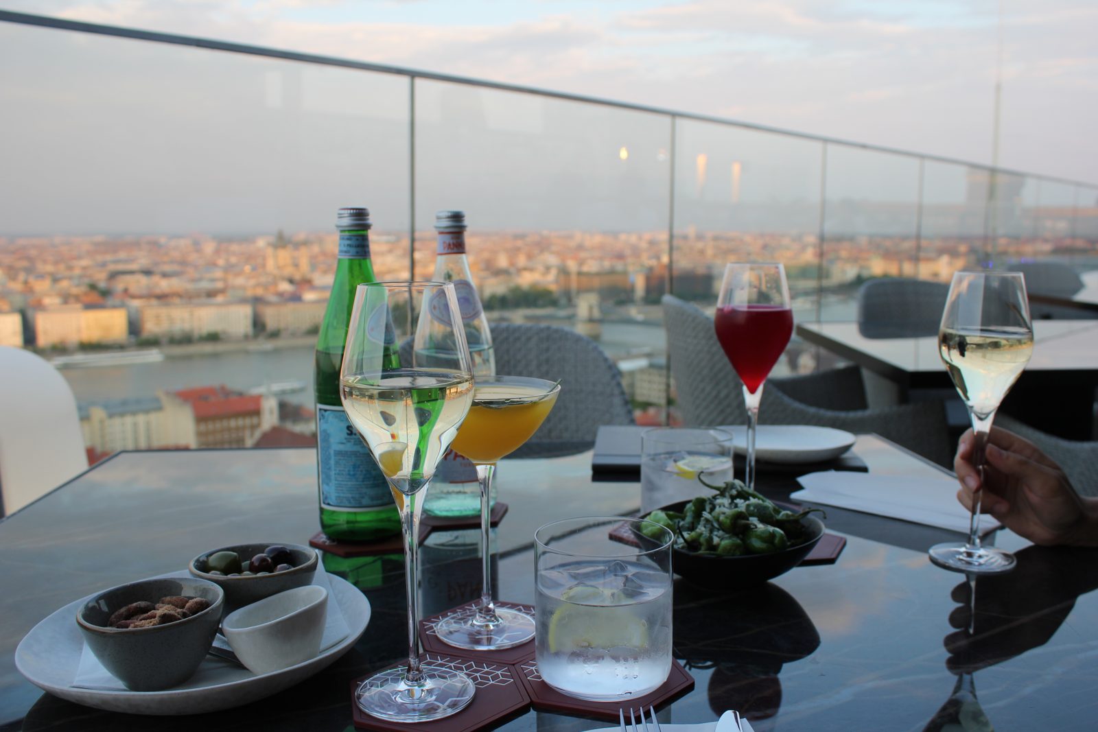 Drinks, olives, nuts and padrón peppers on a table at White Raven Skybar, the Danube and the Pest riverfront behind the rail. Photo: Hungary Unlocked