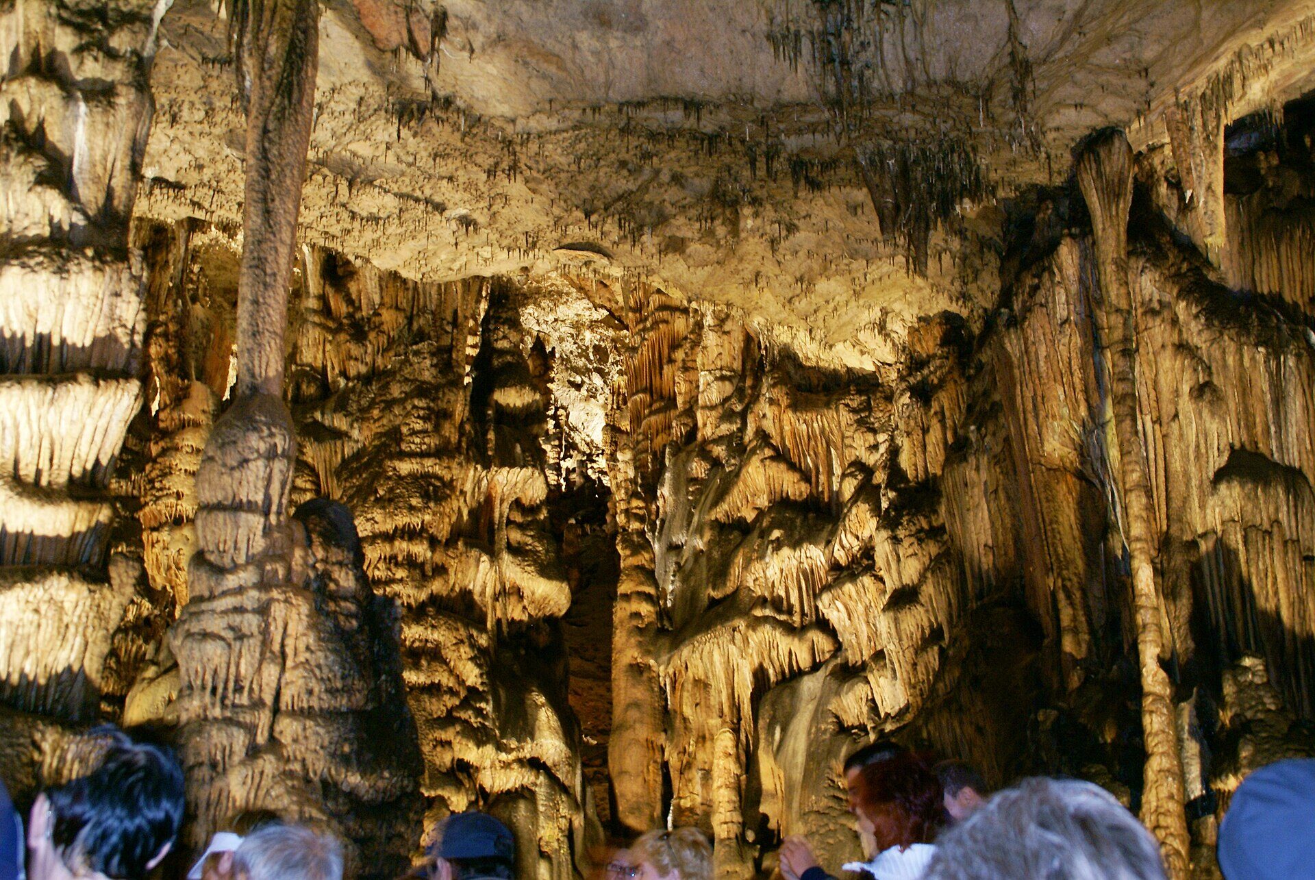 Dense dripstone columns and curtains in the Baradla Cave, lit in warm yellow above the heads of a tour group
