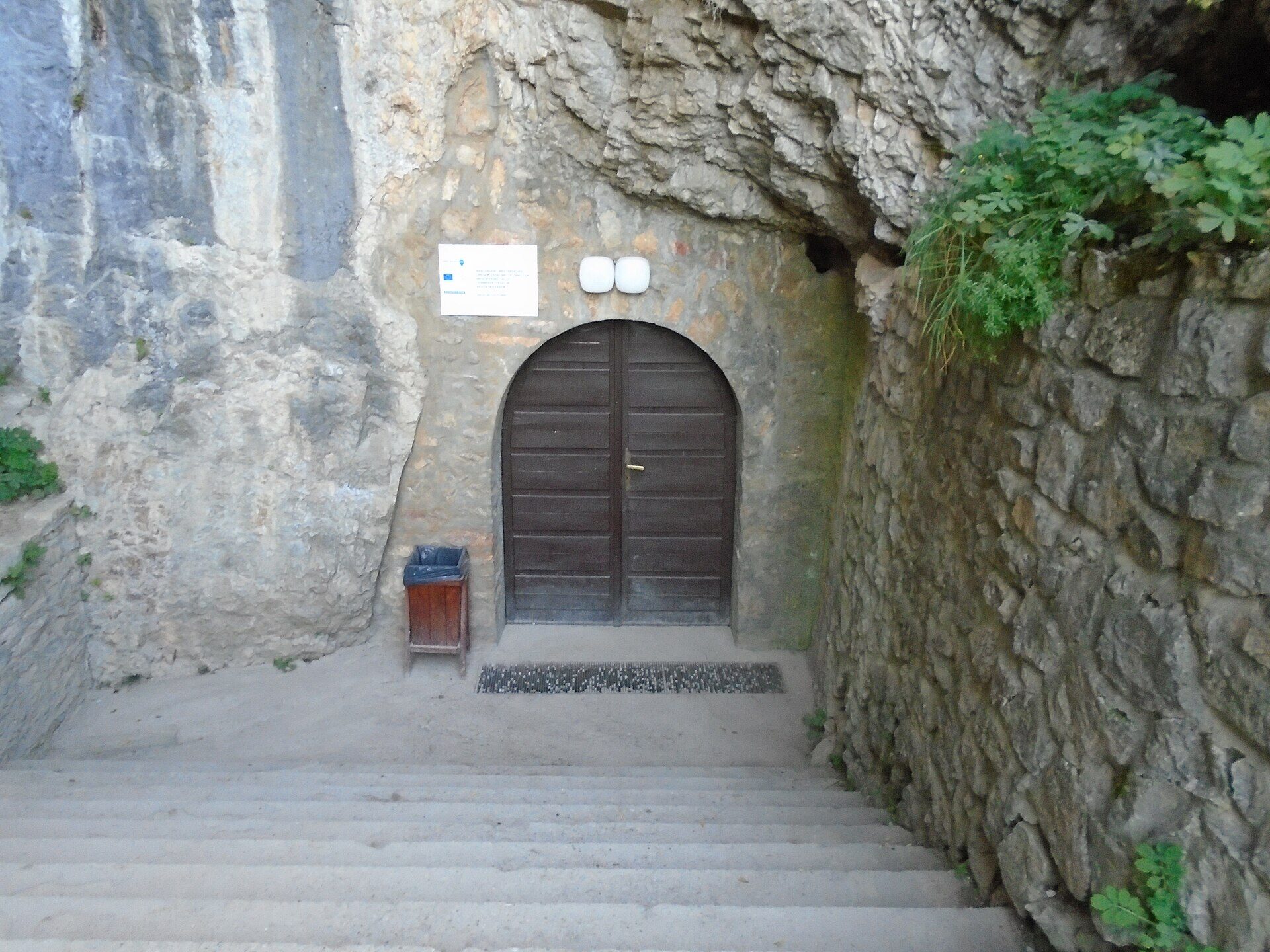 The arched wooden door of the Baradla Cave’s main entrance in Aggtelek, set into a limestone cliff at the bottom of a stone staircase
