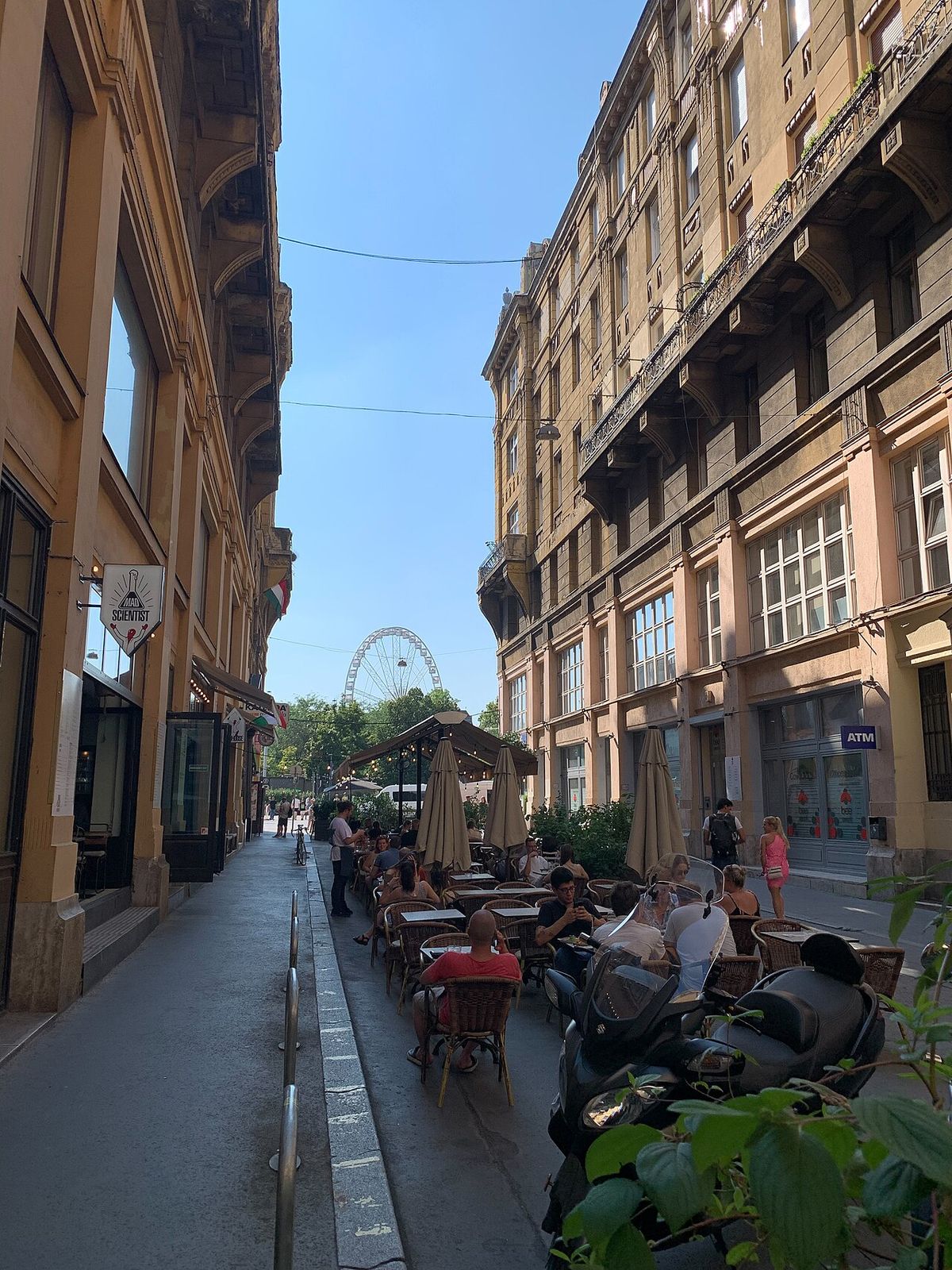 Anker köz in Budapest on a summer afternoon: a narrow lane between tall early-20th-century buildings with café tables and umbrellas along one side and a Ferris wheel visible at the far end