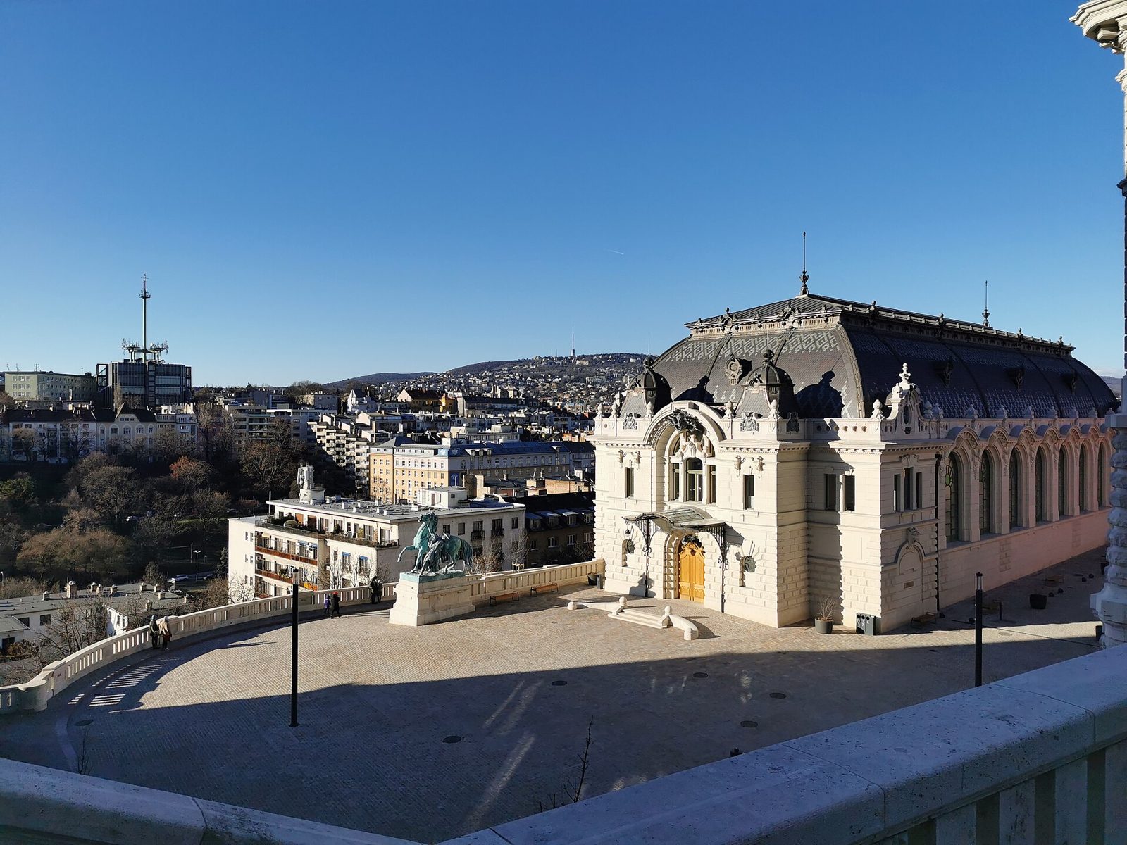 The rebuilt Riding Hall on the Csikós courtyard of Buda Castle, a cream stone hall with a dark curved roof and arched windows, with the bronze horse-tamer statue on the terrace and the Buda hills behind