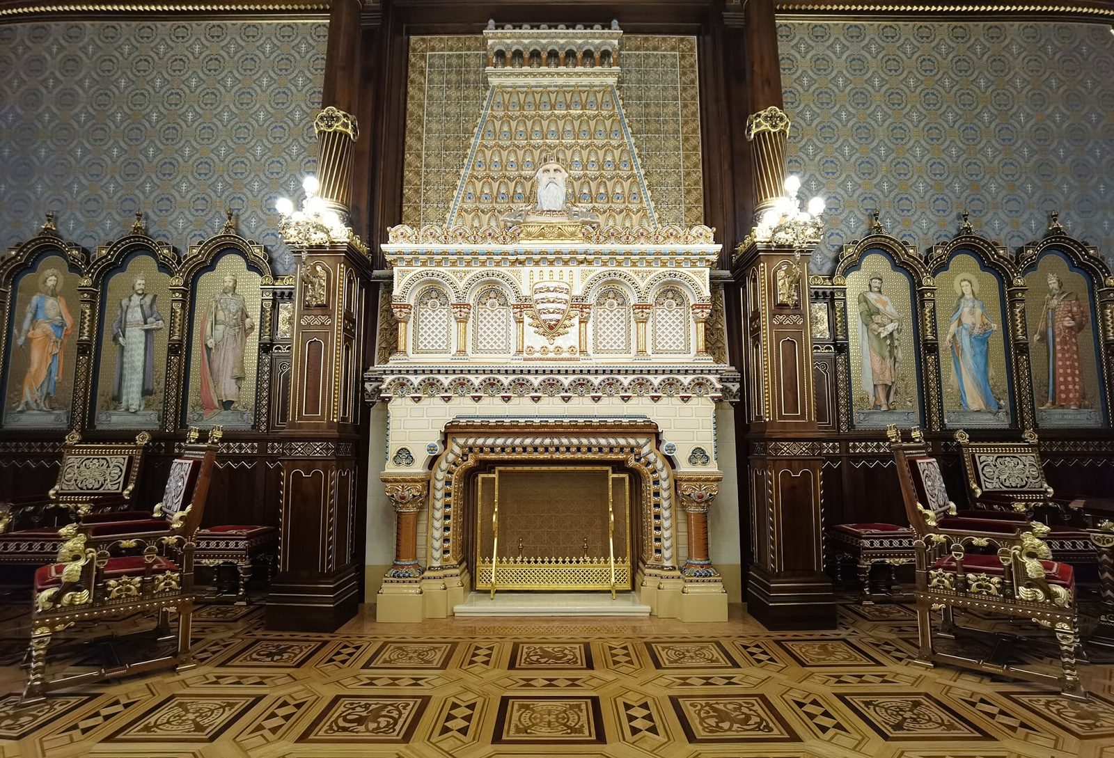 The rebuilt St Stephen’s Hall in Buda Castle, with a white and gold Zsolnay ceramic fireplace between walnut panels holding painted saints, and red-upholstered carved chairs on either side
