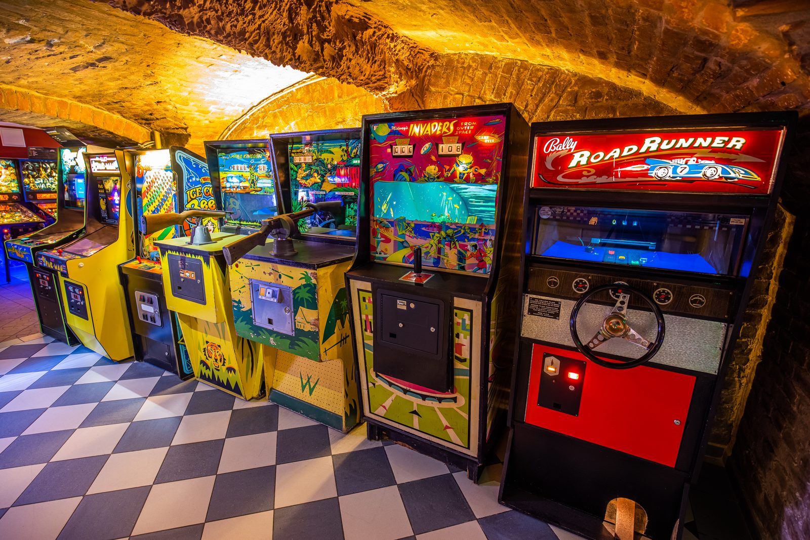 A row of lit vintage arcade cabinets, among them Road Runner and Invaders, under a brick vaulted ceiling at the Budapest Pinball Museum