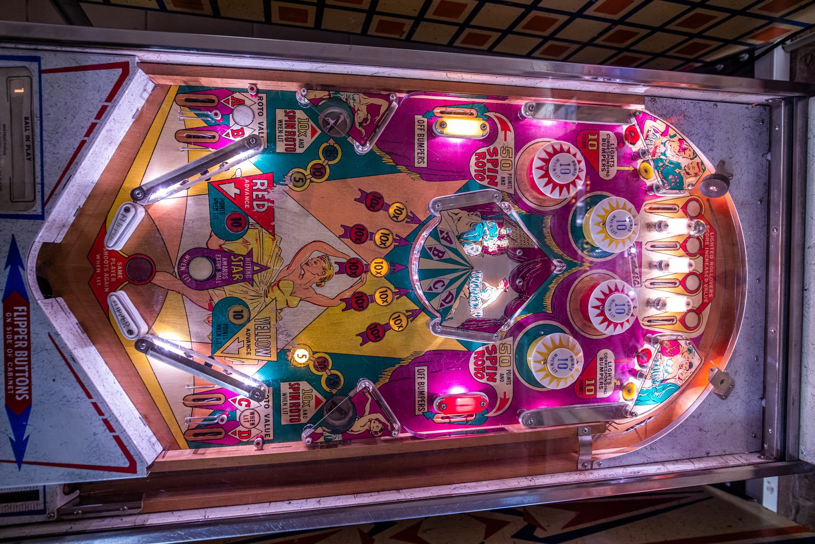 A colourful vintage pinball playfield seen from above, with pop bumpers, flippers and a painted dancer at its centre