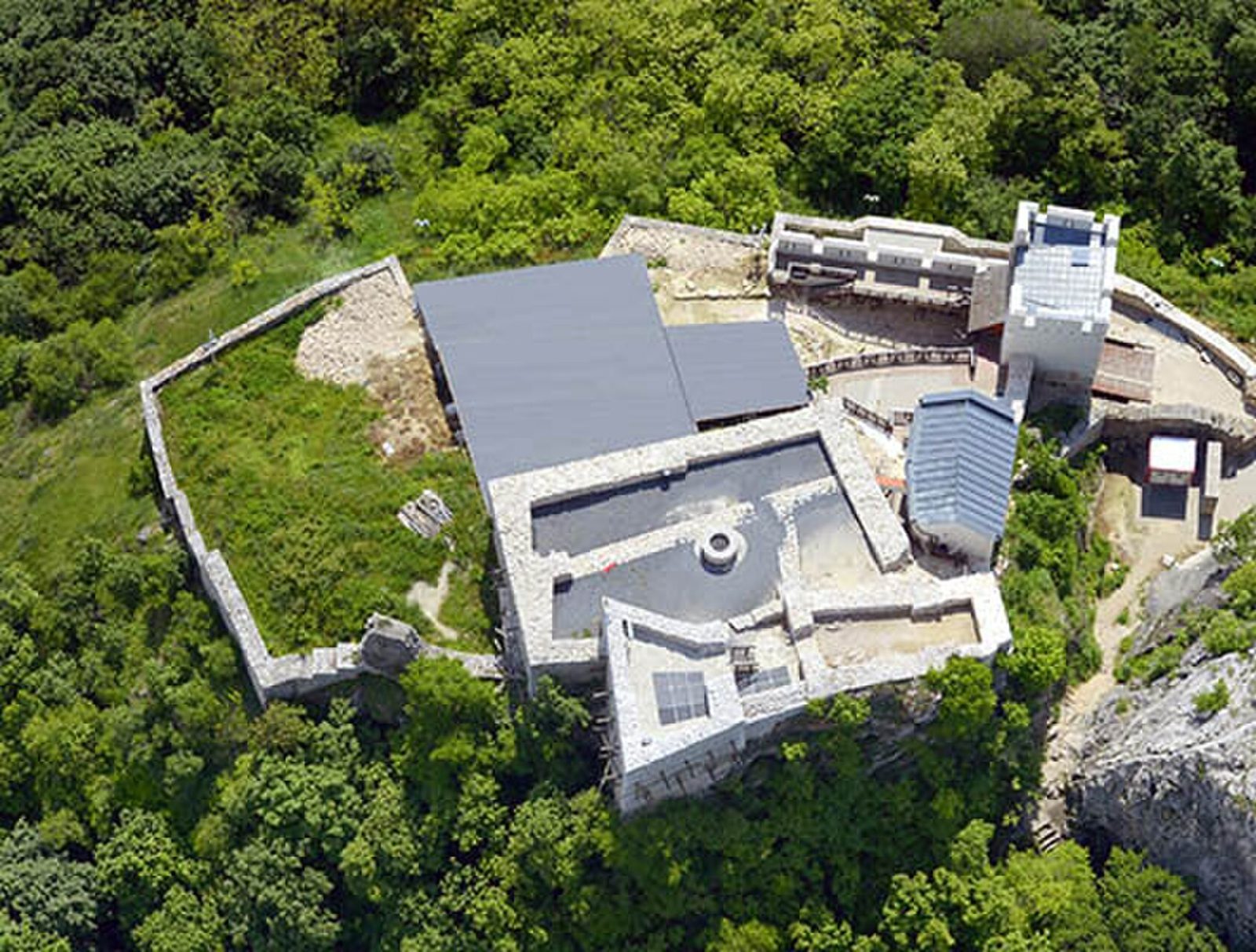 Aerial view of Csókakő Castle on a wooded hilltop, with rebuilt walls, roofed sections and a gate tower above a limestone cliff