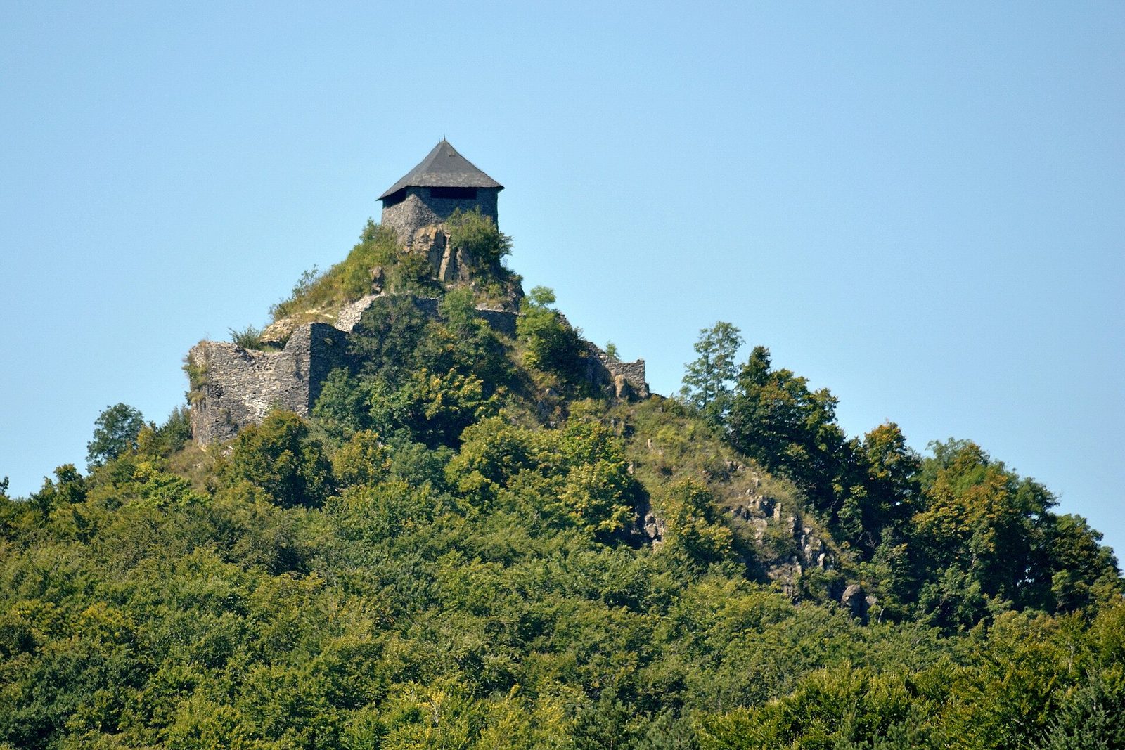 The ruined tower of Salgó Castle with a pointed roof, standing on a rocky, forested volcanic peak under a clear sky